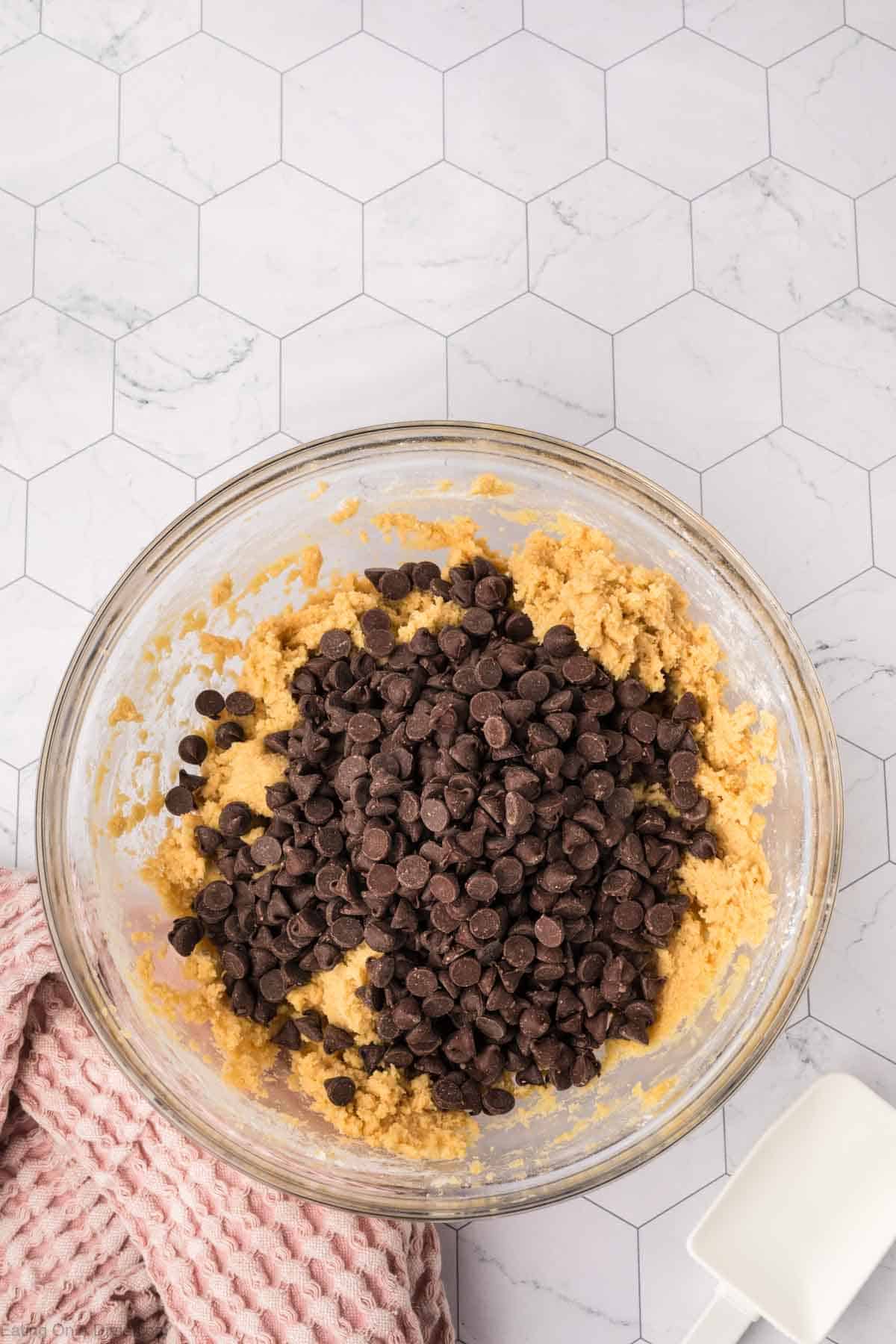 A glass bowl filled with cookie dough, poised for the ultimate giant cookie recipe, is topped with dark chocolate chips on a light, hexagonal-tile counter. A pink textured cloth and a white measuring cup sit nearby.