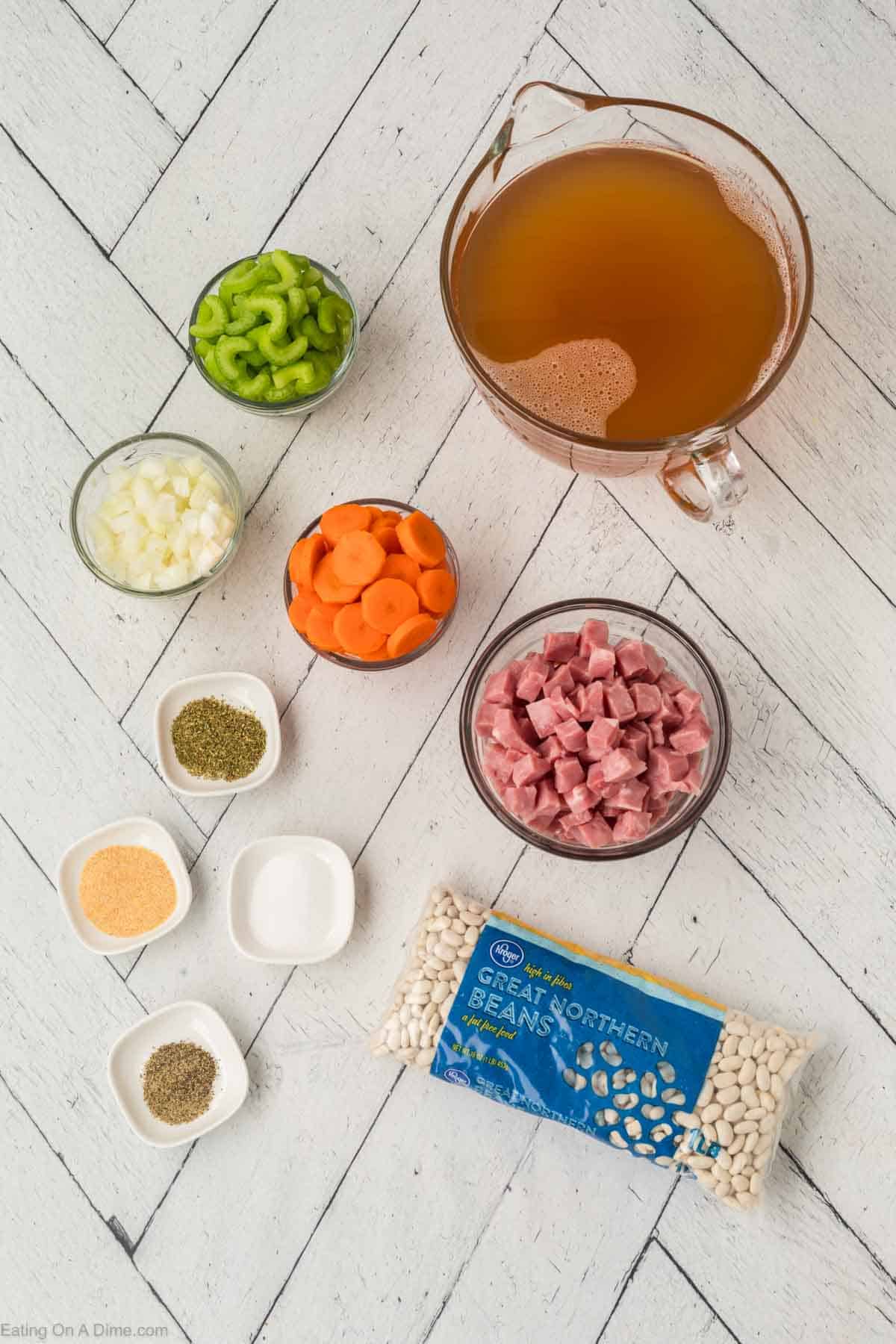 Ingredients for a hearty instant pot ham and bean soup are arranged on a wooden surface, featuring chopped celery, carrots, and onion, diced ham, a bag of Great Northern beans, chicken broth, and small bowls of dried herbs and spices.