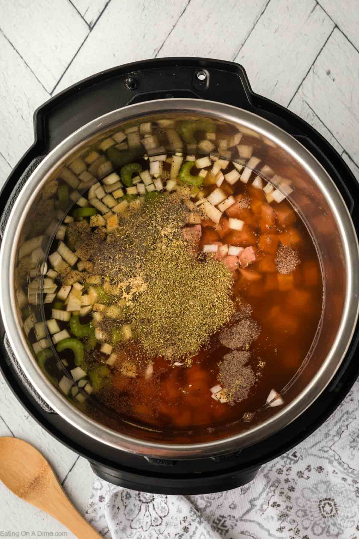 An overhead view of an Instant Pot filled with uncooked ham and bean soup ingredients, including diced onions, celery, meat, and broth. Various spices are sprinkled on top. A wooden spoon and white patterned cloth are nearby on a light-colored surface.