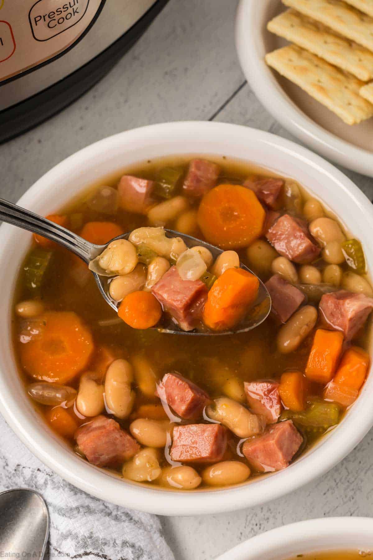 A bowl of instant pot ham and bean soup features chunks of ham, carrot slices, beans, and celery. A spoon lifts the hearty mix. Crackers and a glimpse of the pressure cooker set the cozy scene in the background.