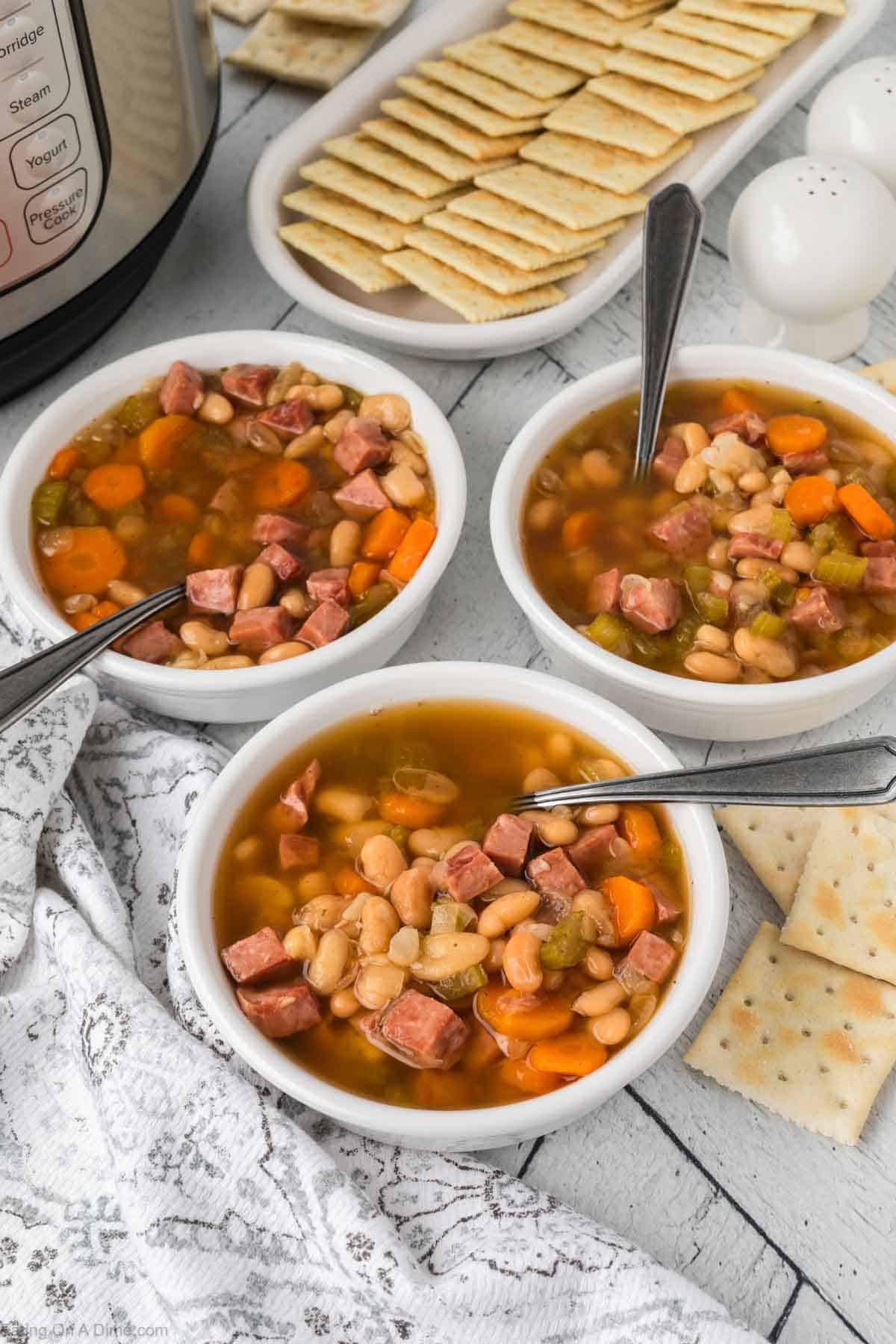 Three bowls of Instant Pot ham and bean soup with carrots and celery are served on a table. Each bowl has a spoon, accompanied by saltine crackers on a plate. The Instant Pot in the background hints at the quick, flavorful preparation, while a patterned cloth adds charm to the setting.