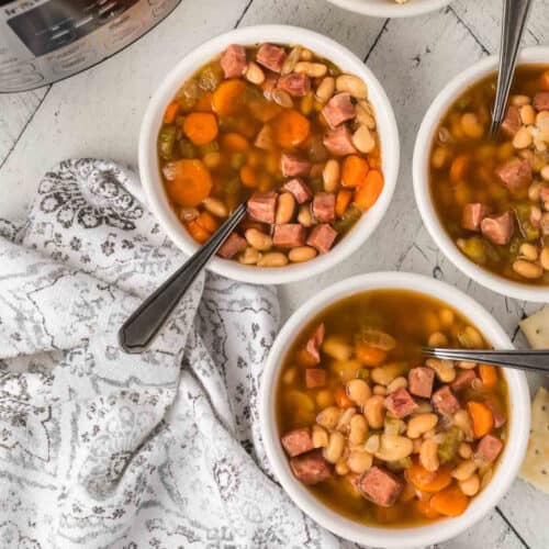 Three bowls of hearty instant pot ham and bean soup sit on a white table, filled with beans, carrots, celery, and chunks of meat. Spoons rest in the bowls, and a patterned cloth lies nearby. A hint of an appliance is visible in the background.