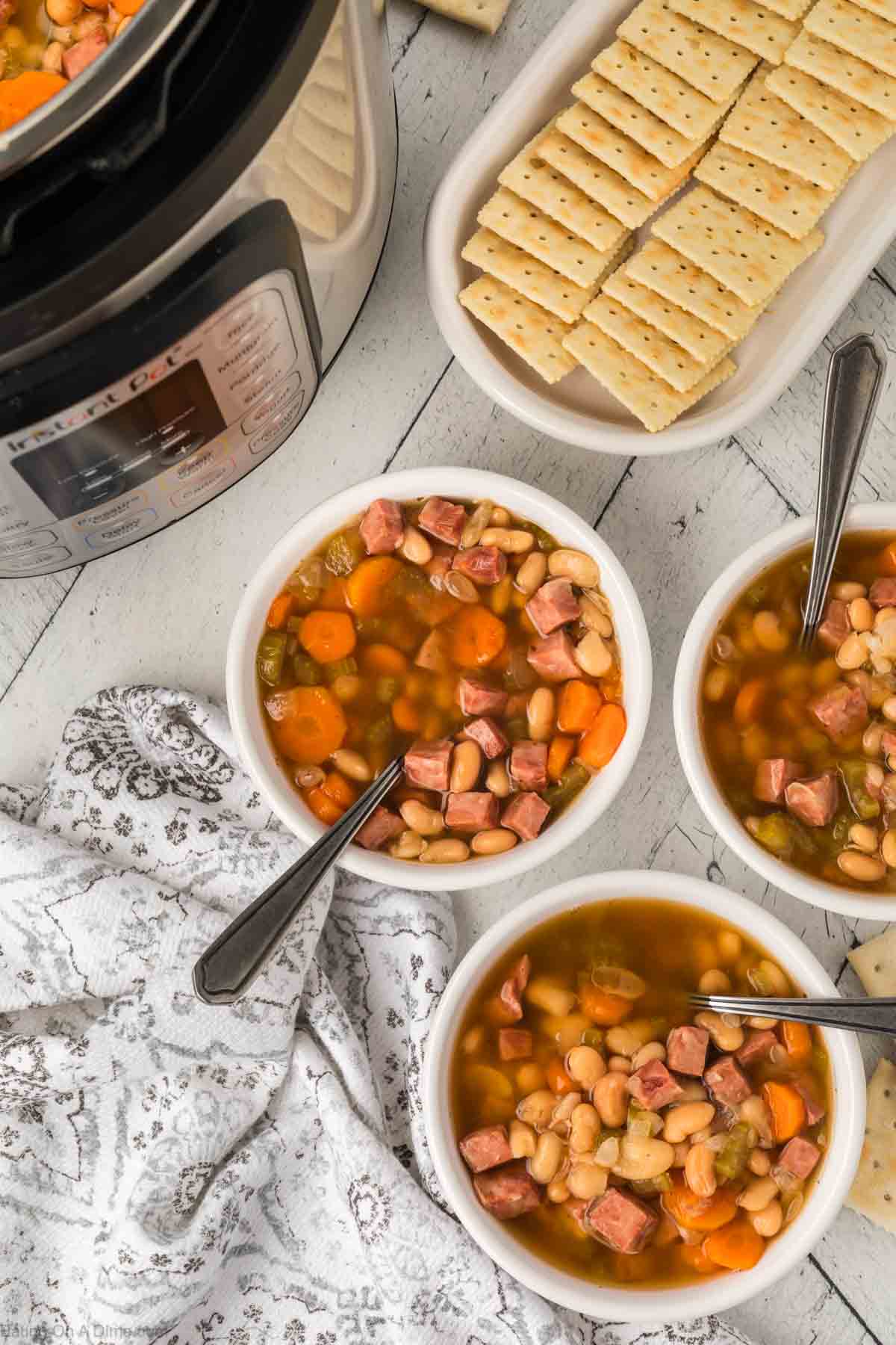 Bowls of Instant Pot ham and bean soup with carrots and celery are served with spoons. A tray of crackers rests beside the pressure cooker on a rustic wooden table, alongside a patterned cloth.