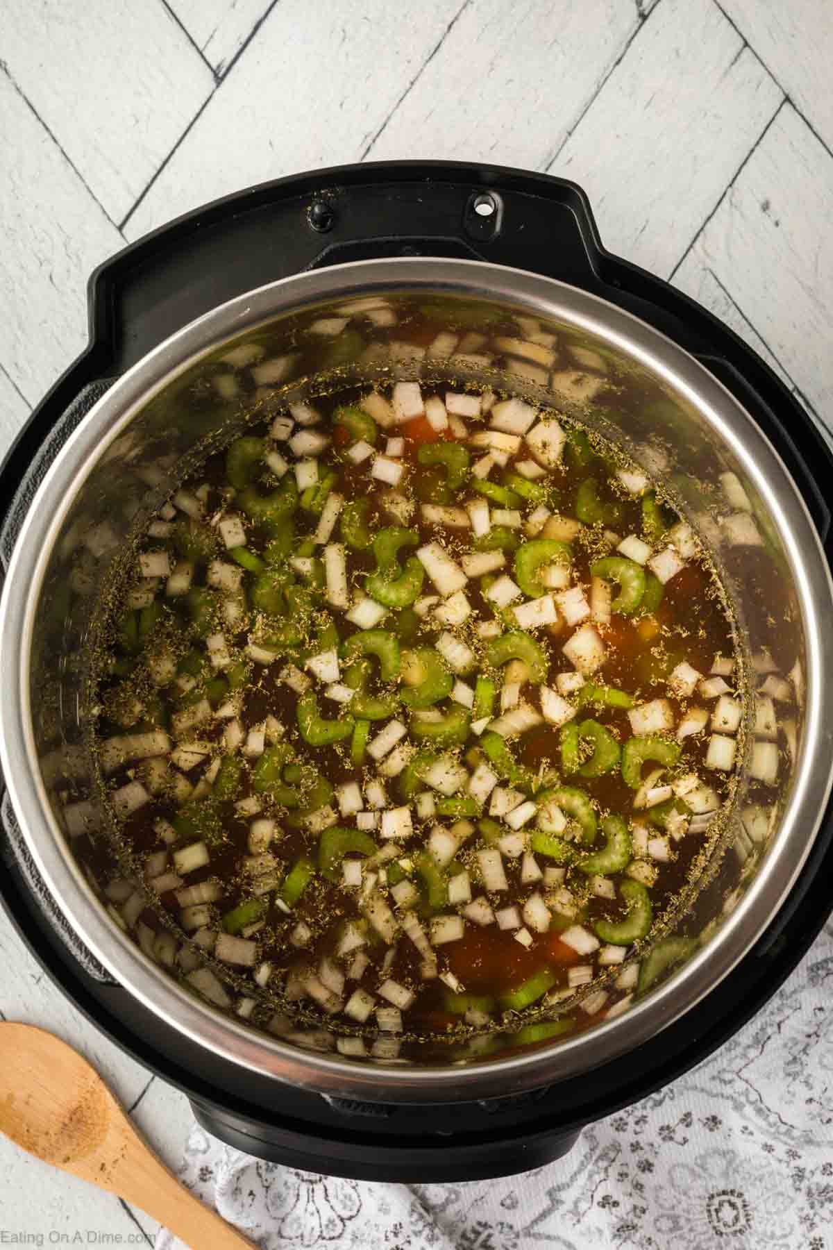 A top view of an Instant Pot filled with chopped vegetables and tender ingredients for a hearty ham and bean soup, including celery and onions in a rich brown broth. The pot rests on a light-colored wooden floor with a patterned cloth and wooden spoon nearby.