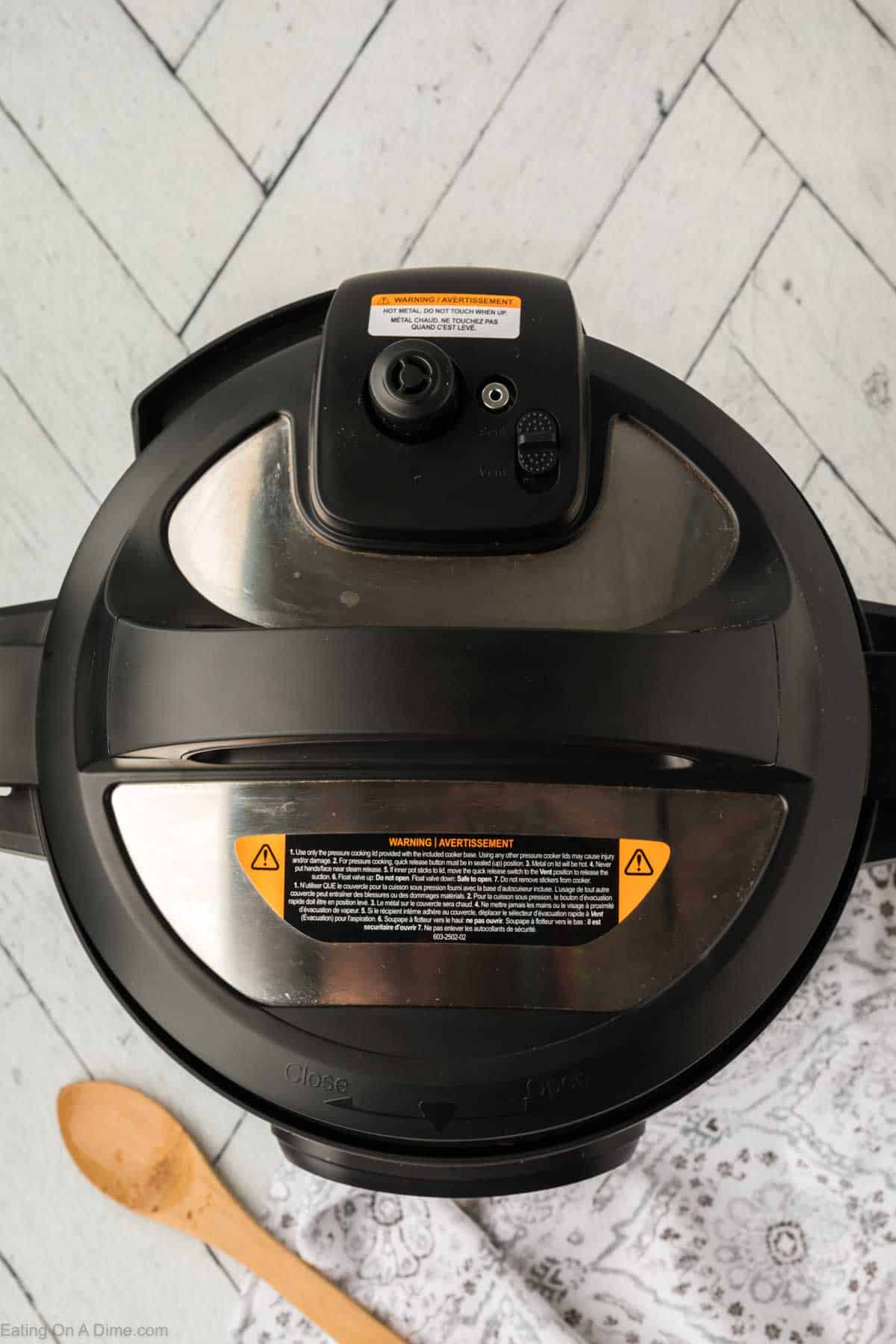 Top view of a black and silver multi-cooker on a light wood floor, perfect for whipping up an instant pot ham and bean soup. A wooden spoon and patterned white cloth sit beside it, while warning labels and control buttons grace the lid.