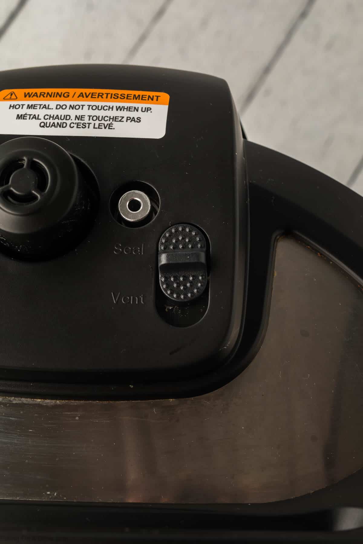 Close-up of a pressure cooker lid, perfect for making instant pot ham and bean soup, featuring a black switch with "Seal" and "Vent" options. A warning label in English and French cautions not to touch when hot. The reflective lid has a textured grip for safety.