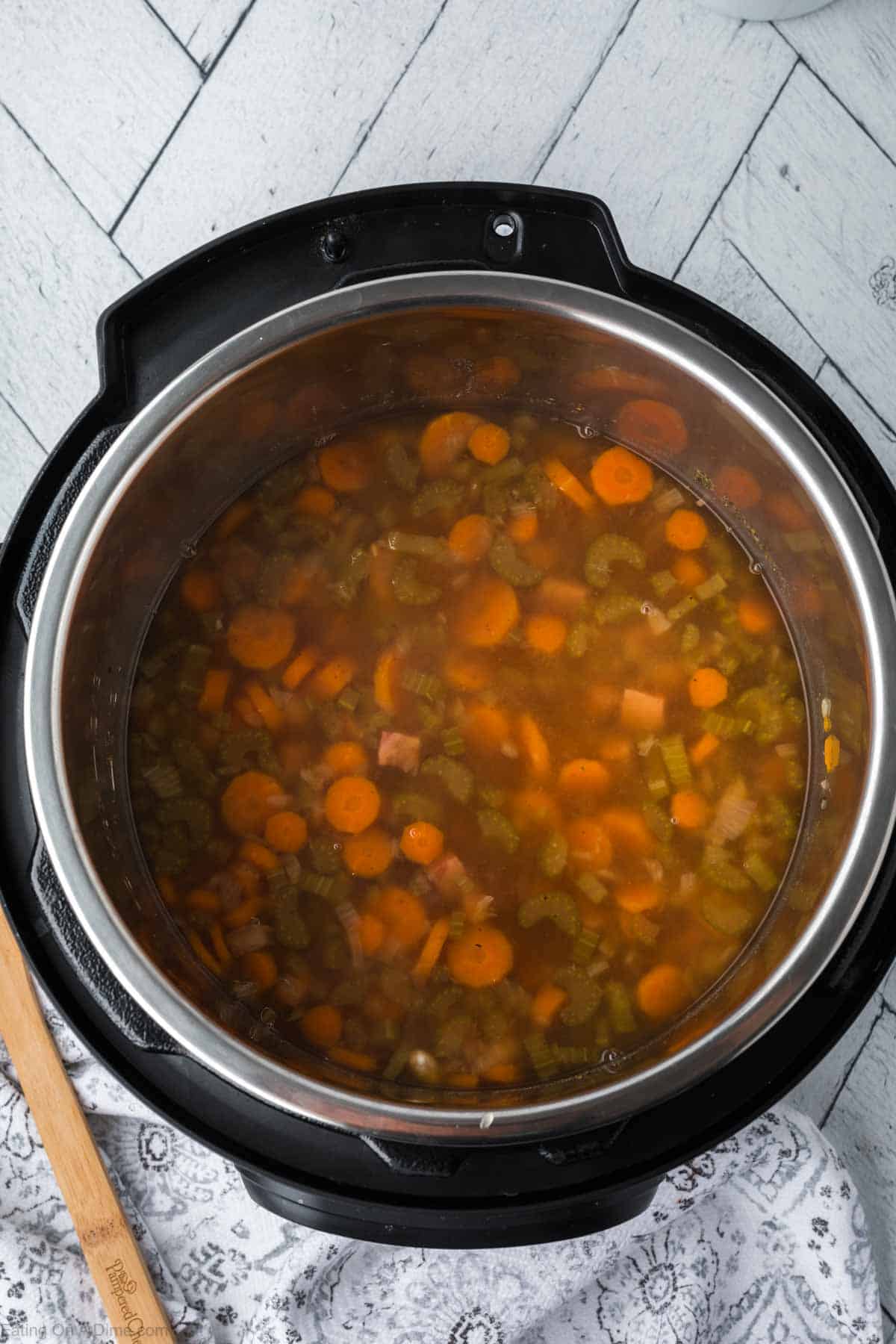 A pot of instant pot ham and bean soup, enriched with slices of carrots, celery, and other vegetables in a clear broth, sits on a patterned cloth placed on light wooden flooring. A wooden spoon rests against the pot.