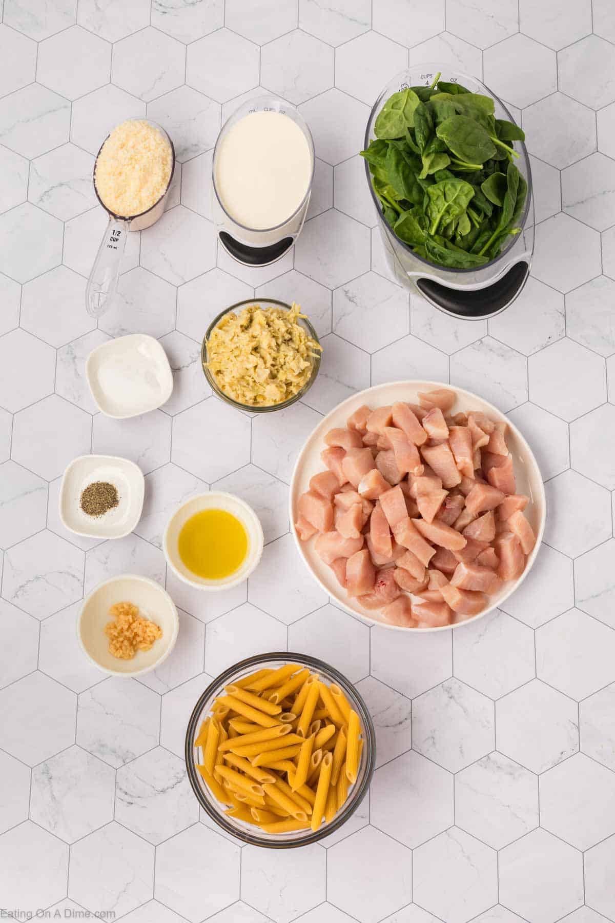 A top view of various ingredients arranged on a light gray geometric background. Ingredients include chopped raw chicken in a bowl, spinach, uncooked penne pasta, minced garlic, grated cheese, oil, spices, artichoke dip for Chicken Spinach Artichoke Pasta, and a container of cream.