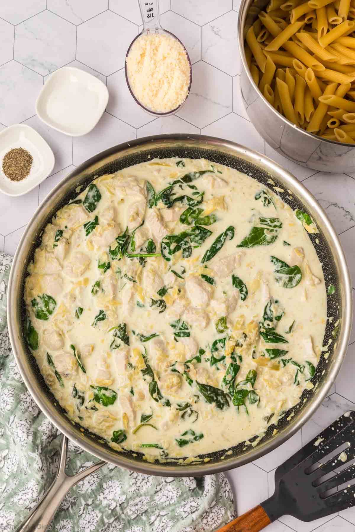 A skillet filled with creamy Chicken Spinach Artichoke Pasta on a white hexagonal tile surface. Surrounding the skillet are uncooked pasta in a pot, a bowl of grated cheese, a small dish of pepper, another small dish with sauce, and a cloth napkin.