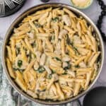 A close-up of a pan filled with creamy chicken spinach artichoke penne pasta. The dish features penne pasta mixed with chunks of chicken, fresh spinach, and artichokes, all coated in a luscious sauce. A small bowl of grated cheese is visible in the background alongside cutlery and plates.