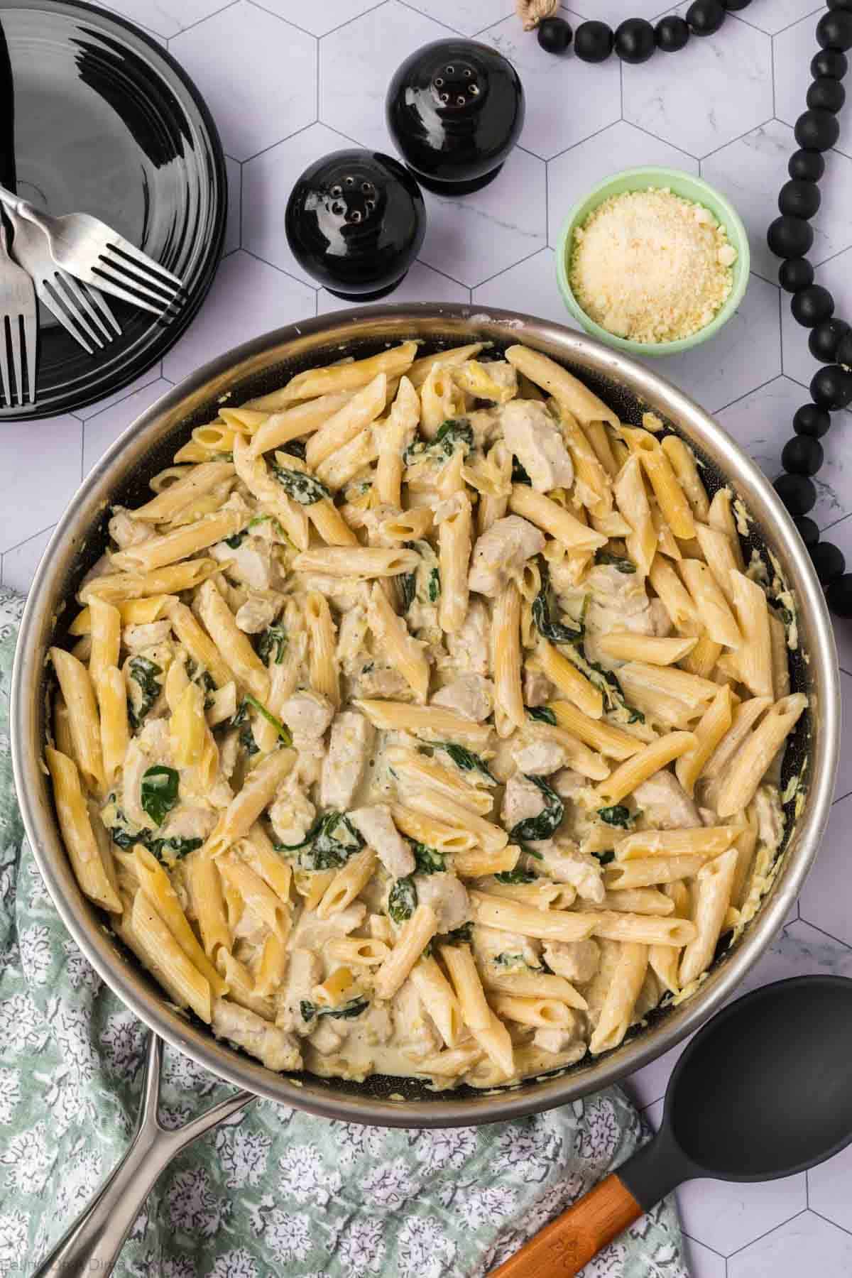 A skillet filled with creamy Chicken Spinach Artichoke Pasta is on a hexagon-patterned table. Surrounding the skillet are black pepper and salt shakers, a bowl of grated cheese, a black ladle, and a stack of black plates with forks on a patterned cloth napkin.