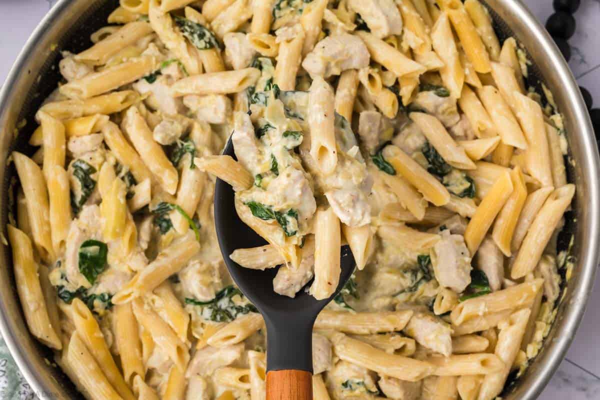 A pan filled with creamy Chicken Spinach Artichoke Pasta, showing penne noodles, pieces of chicken, and bits of spinach. In the center, a black spoon with a wooden handle is holding a serving of the pasta.