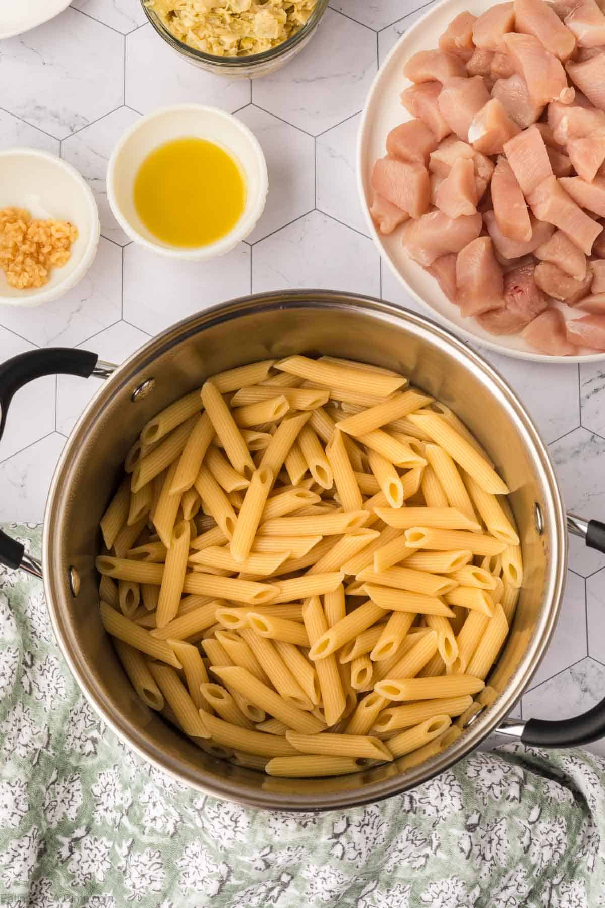 A cooking preparation scene shows a pot filled with cooked penne pasta on a countertop. Surrounding the pot are small bowls containing minced garlic, olive oil, a creamy sauce, and a plate of diced raw chicken. A patterned cloth partially covers the counter, setting the stage for Chicken Spinach Artichoke Pasta.