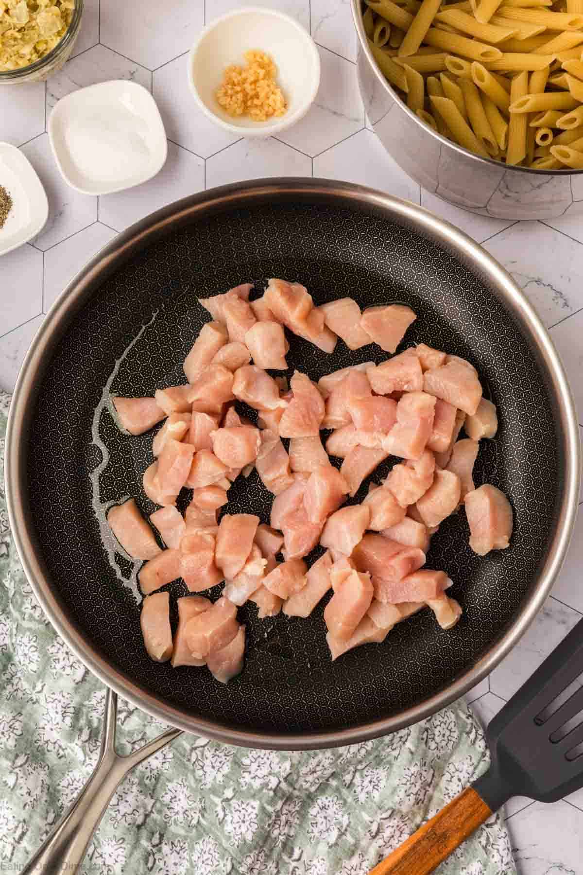 Chicken pieces are cooking in a black frying pan on a stovetop. Surrounding the pan, there are small bowls containing minced garlic, seasonings, and a pot of uncooked penne pasta. A spatula with a wooden handle rests nearby on a green and white patterned cloth, ready to stir the Chicken Spinach Artichoke Pasta masterpiece.