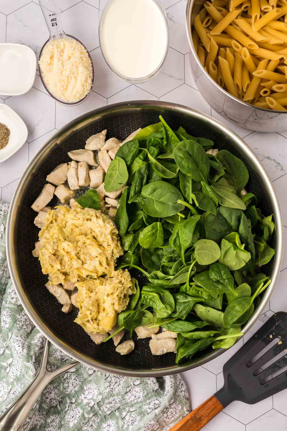 A skillet containing a mixture of cooked chicken, fresh spinach, and artichoke dip. Surrounding the skillet are ingredients for Chicken Spinach Artichoke Pasta: dry penne pasta in a pot, grated parmesan cheese, a small white bowl with a spice, cream, and a patterned cloth.