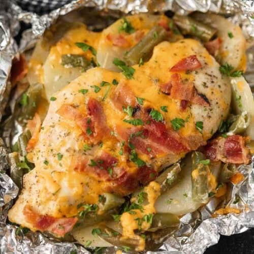 A Bacon Ranch Chicken Foil Packet features a baked chicken breast topped with melted cheese, cooked bacon pieces, and a dash of parsley. This delicious meal is complemented by sliced potatoes and green beans, all conveniently wrapped in foil for an easy-to-prepare feast.