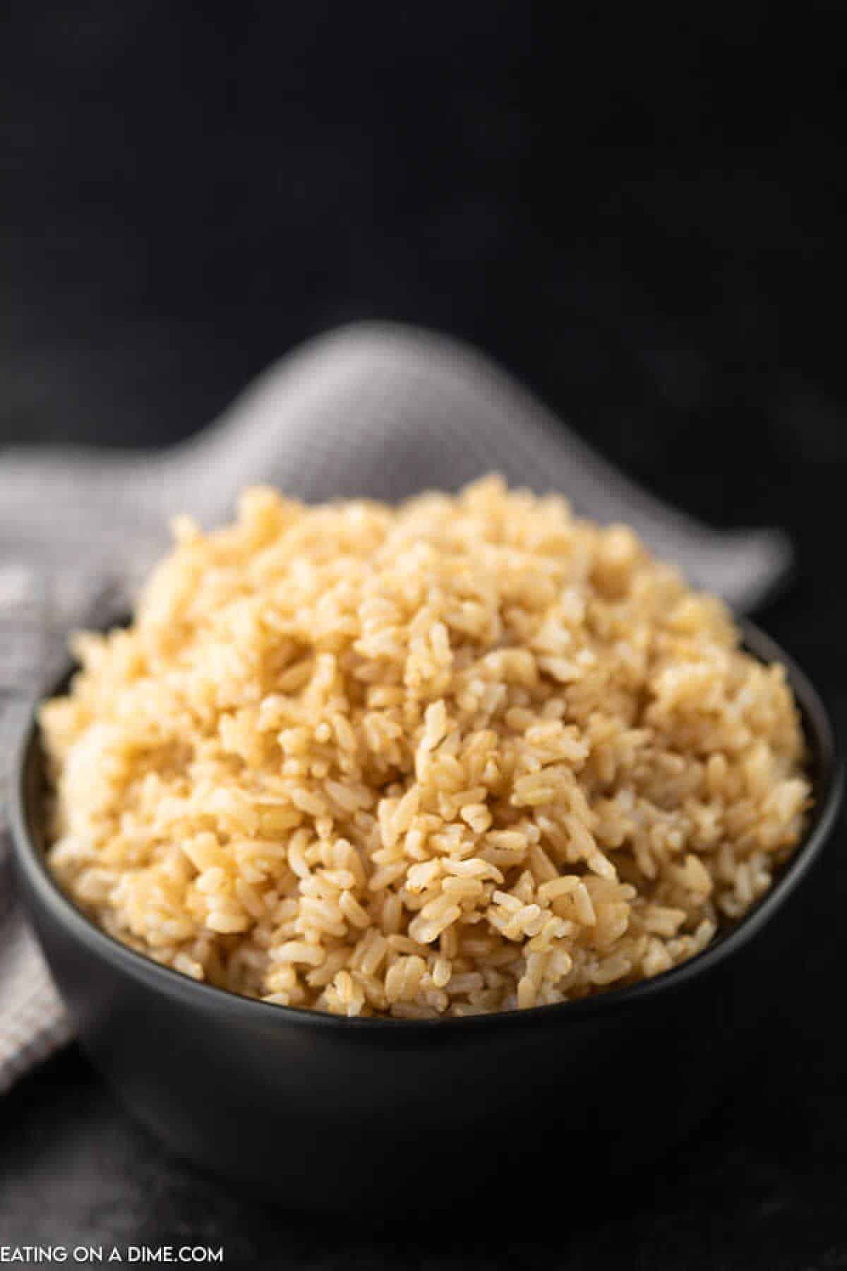 A bowl of Instant Pot brown rice is placed on a dark surface, with a textured gray cloth in the background. The rice appears well-cooked, with a light and fluffy texture.