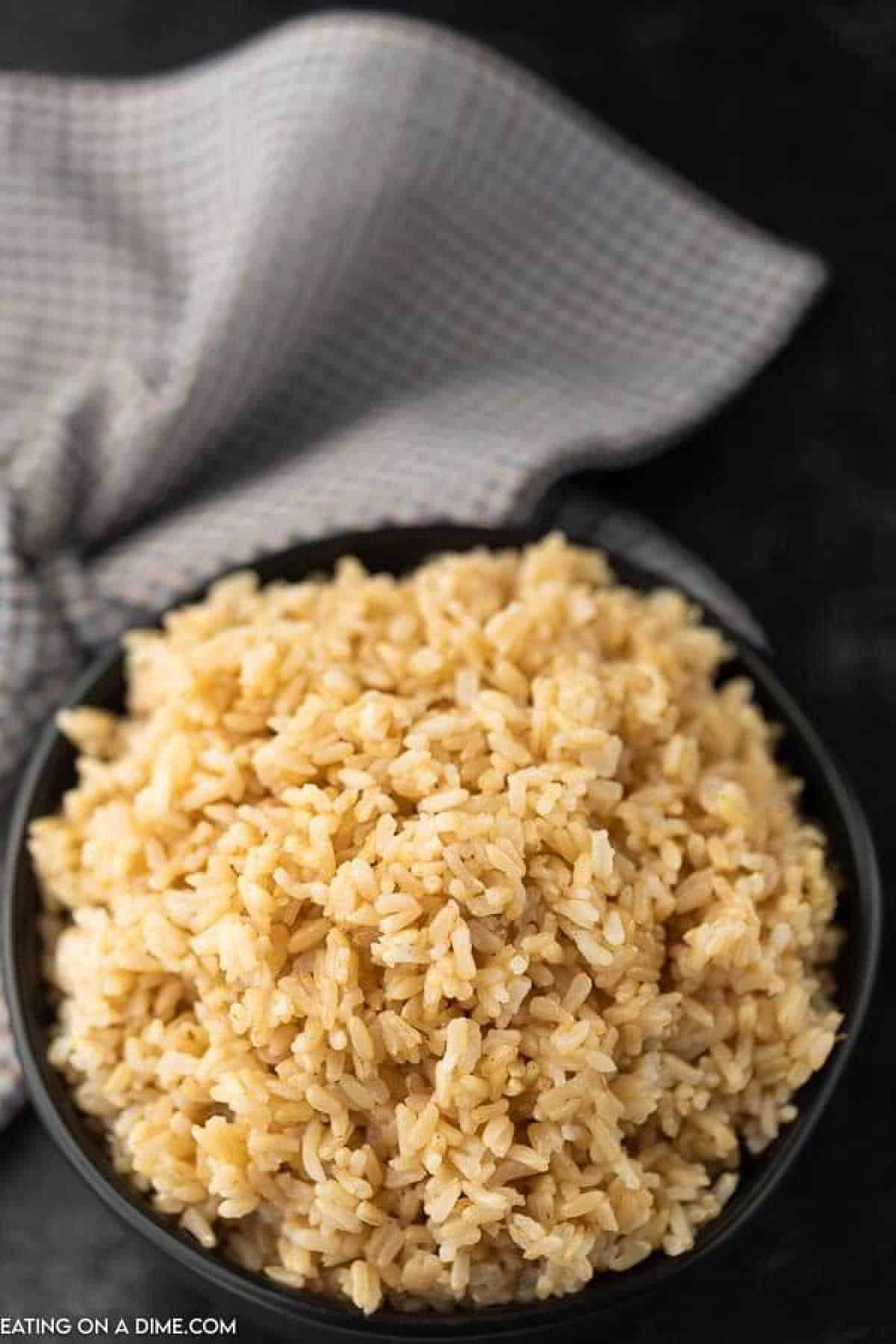 A bowl filled with Instant Pot brown rice is placed on a dark surface. A textured gray cloth is casually draped in the background.