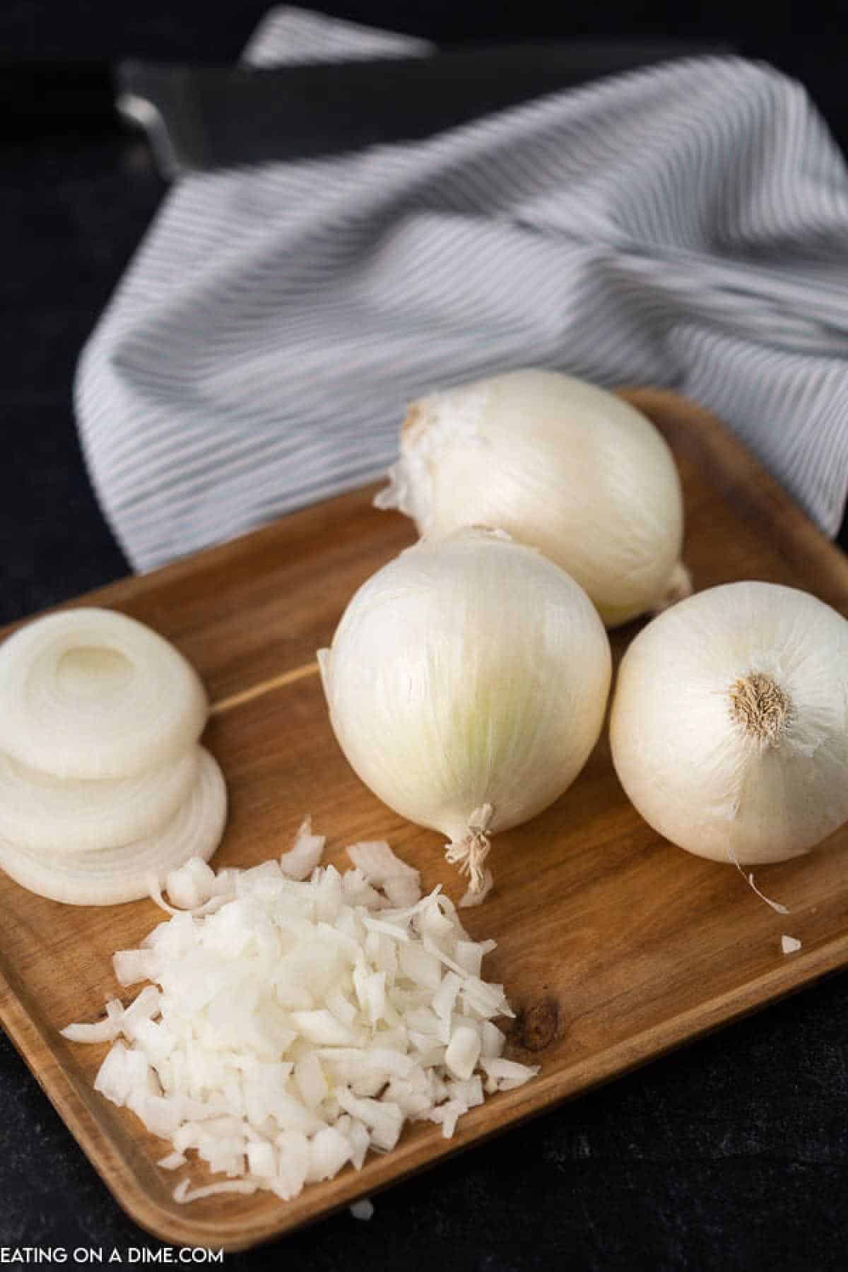 A wooden tray holds three whole white onions, a few sliced onion rings, and a pile of chopped onions. Learn how to cut onions without crying as a striped cloth and knife lie ready in the background on the dark surface.