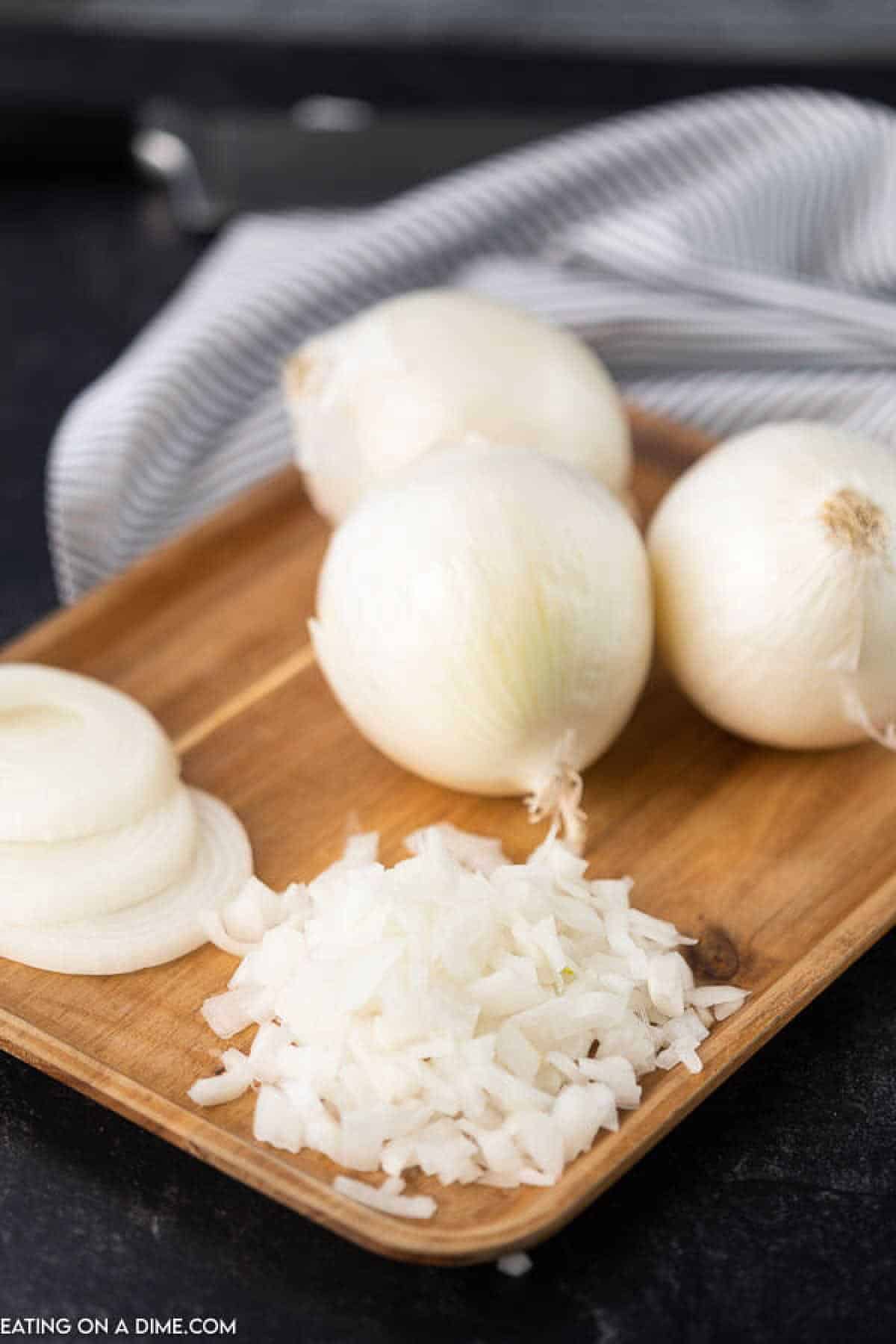 A wooden tray displays three whole white onions, a few slices, and a small pile of finely chopped bits. A striped cloth is partially visible in the background, hinting at tips on how to cut onions without crying.