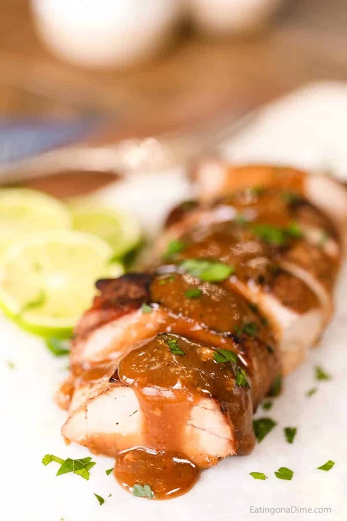 Sliced grilled chicken breast drizzled with a brown sauce, garnished with chopped herbs. Lemon slices add a touch of color to the wooden table setting, reminiscent of a honey lime zest. A knife is partially visible at the top.