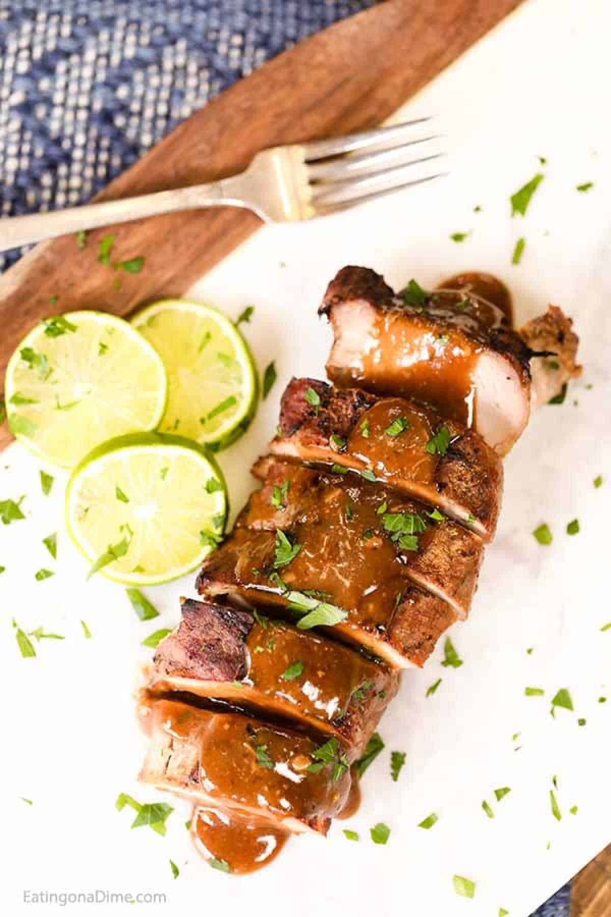 Sliced grilled honey lime pork tenderloin topped with brown sauce and garnished with chopped herbs, served on a white plate. Lime slices are placed next to the meat, and a fork rests nearby on a wooden surface, inspired by our crock pot recipe.