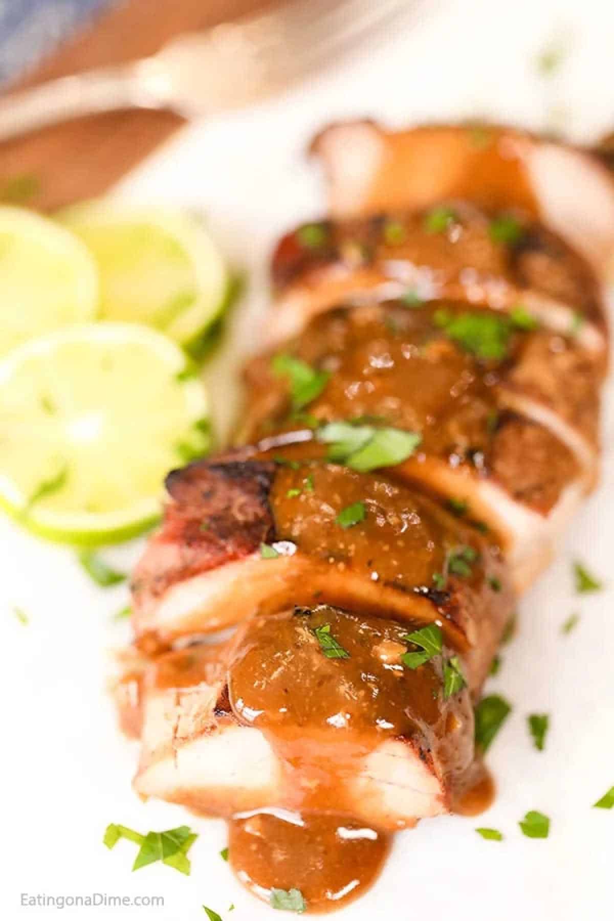 Sliced grilled pork tenderloin topped with a rich brown sauce and garnished with chopped herbs. Lime slices accompany the meat on a white plate, adding a zesty twist to this delightful honey lime crock pot pork tenderloin recipe.