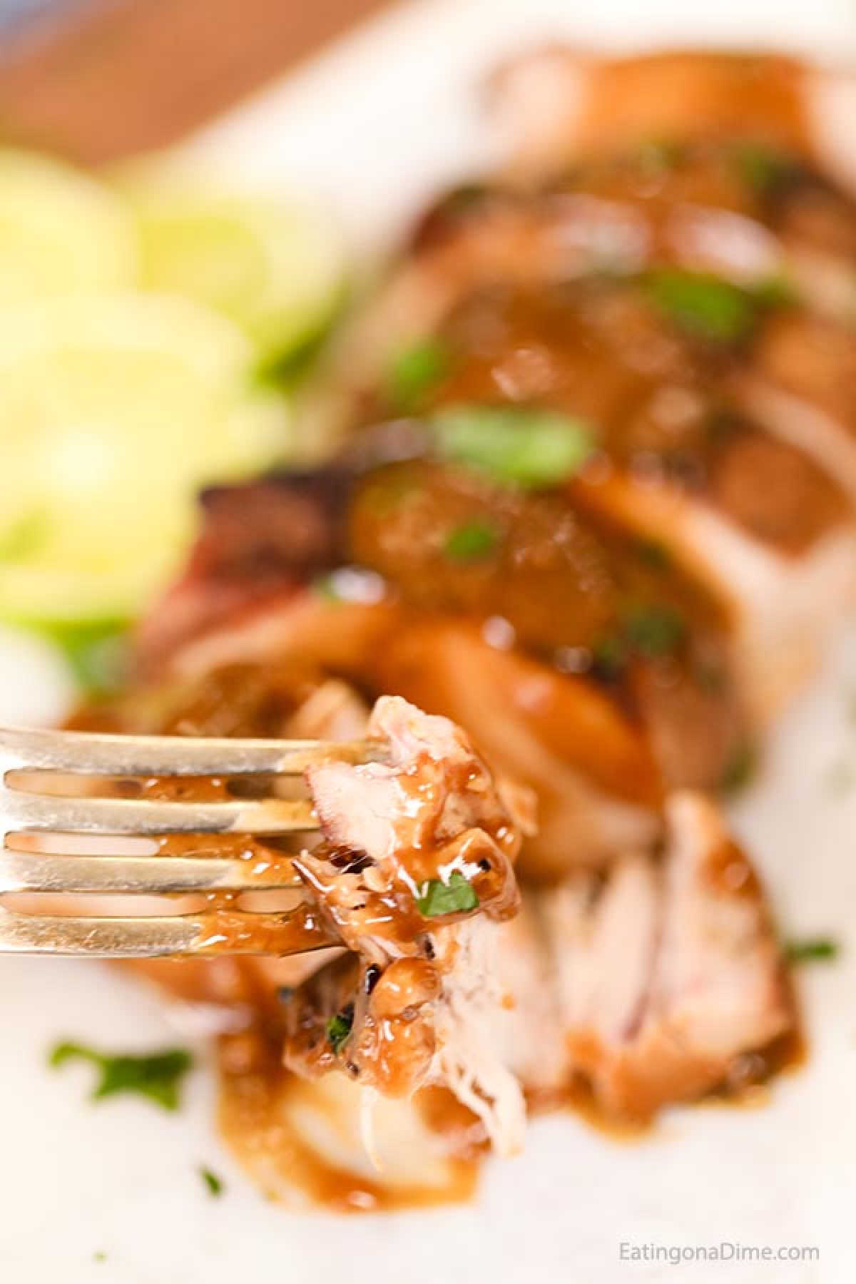 A fork holds a piece of glazed chicken with herbs, positioned in front of a sliced roasted chicken breast on a plate. Echoing the flavors of a Honey Lime Crock Pot Pork Tenderloin recipe, slices of lime are artfully arranged in the background.