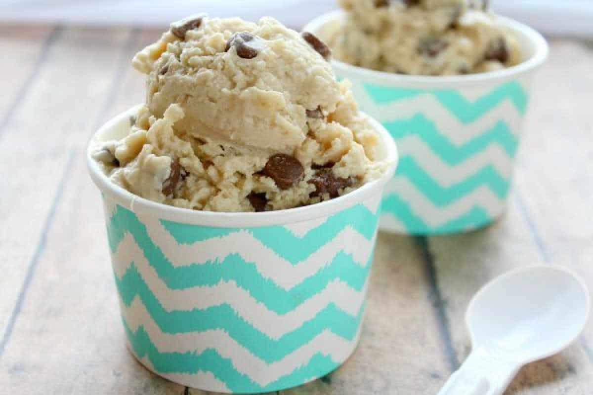 Two cups of chocolate chip cookie dough frozen yogurt are in zigzag-patterned containers on a wooden surface, with a white plastic spoon beside them.