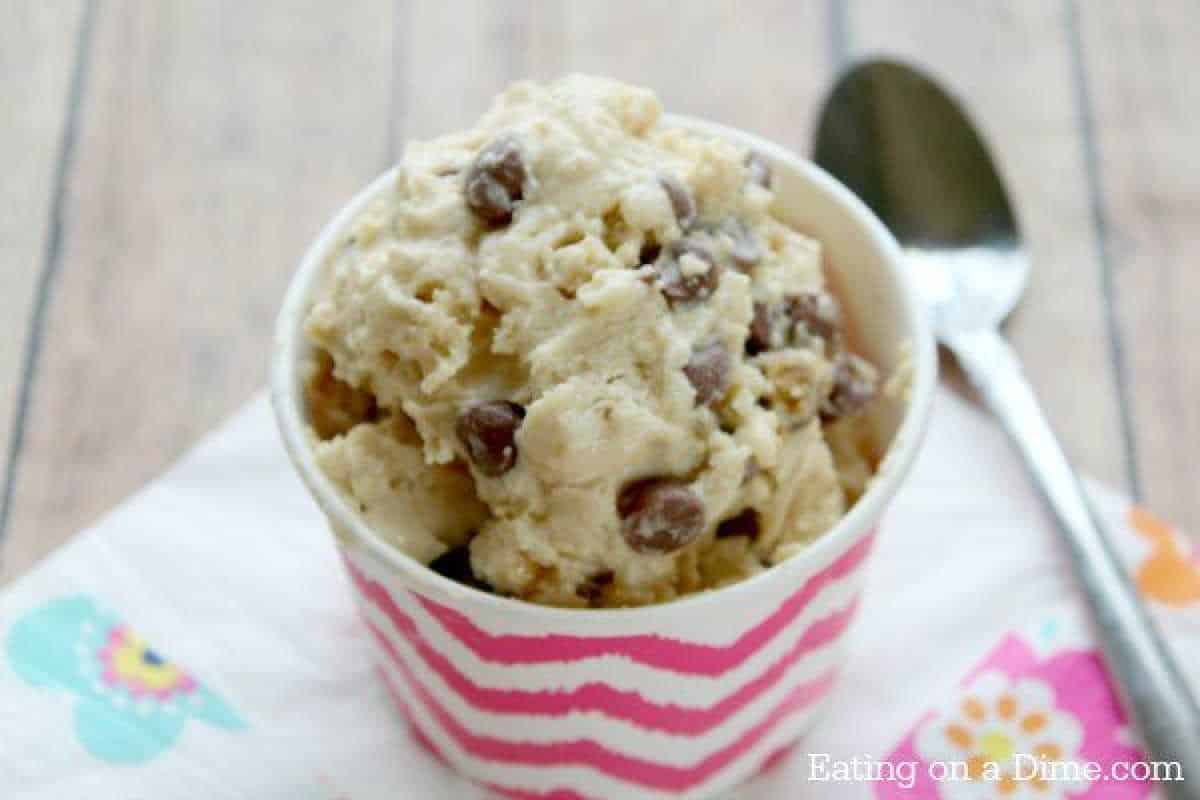 A cup filled with chocolate chip cookie dough frozen yogurt sits on a vibrant floral napkin. A silver spoon is placed next to the cup on a wooden surface. The cup has pink and white zigzag patterns, adding a playful touch to this delightful treat.