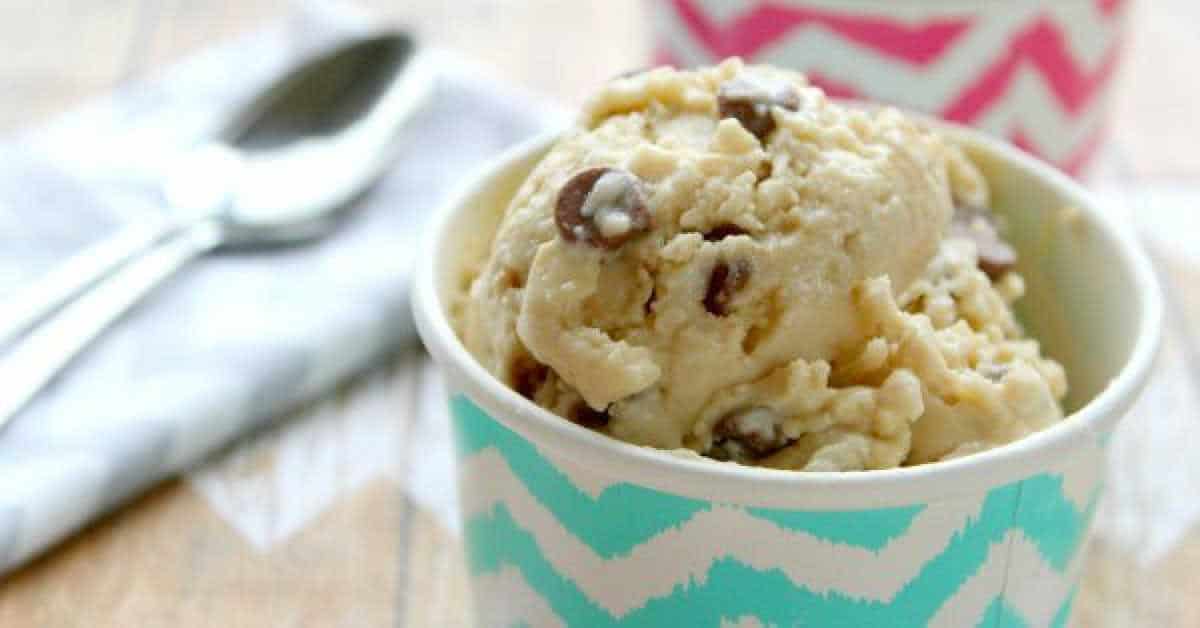 A close-up of a scoop of chocolate chip cookie dough frozen yogurt glistens with chocolate chips in a blue and white zigzag patterned cup. In the background, there's another cup with a pink and white pattern, alongside a napkin and spoon.