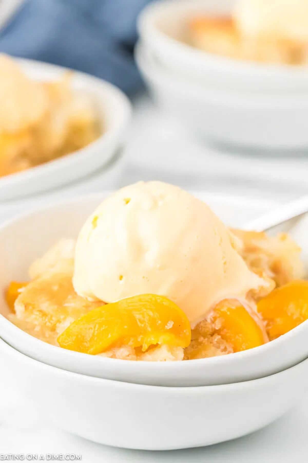 A delicious crock pot peach cobbler topped with a scoop of vanilla ice cream sits invitingly in a white bowl. The dessert showcases juicy peach slices and a crumbly texture, while a spoon is partially visible beside the bowl, ready for you to savor each bite.