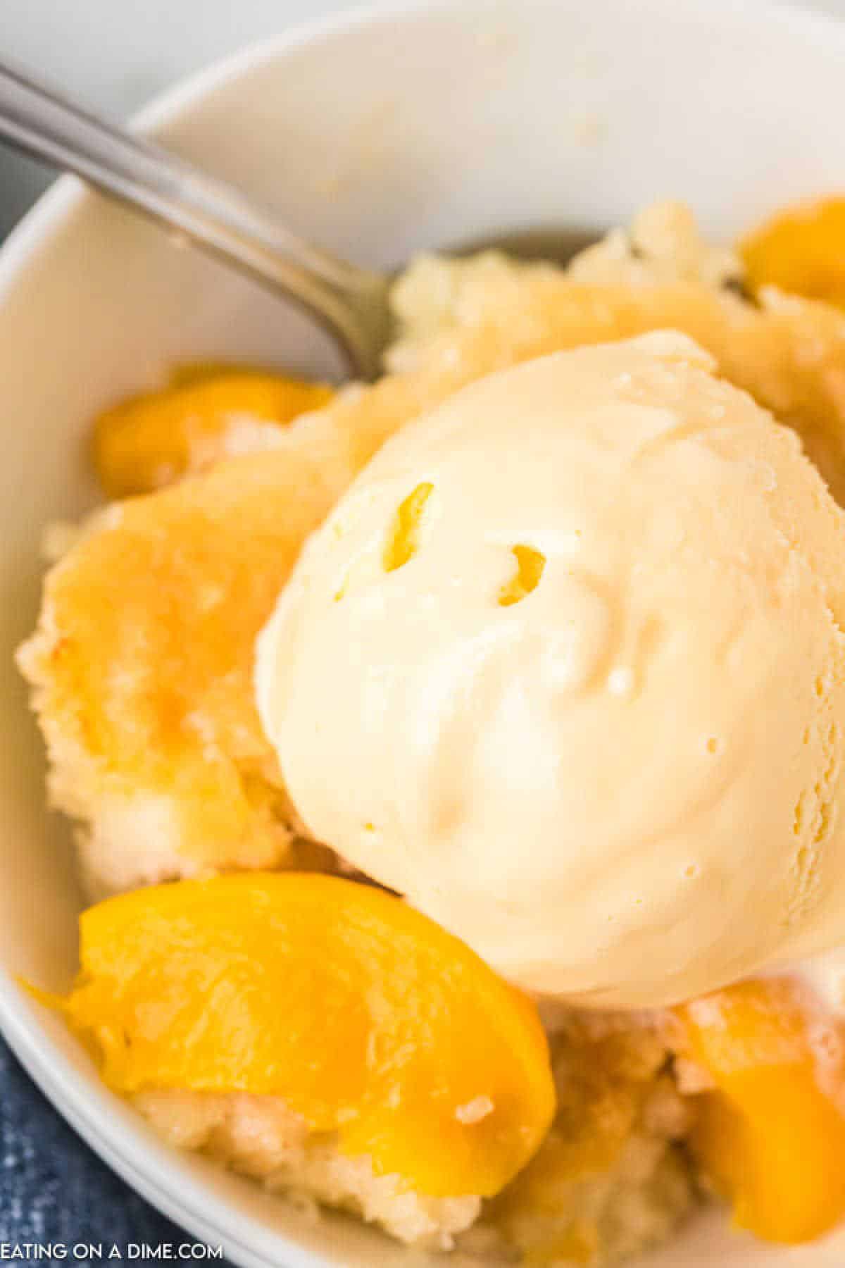 A close-up of a dessert bowl featuring a scoop of vanilla ice cream on top of crock pot peach cobbler. The cobbler has visible peach slices and a golden crust, with a spoon in the background.