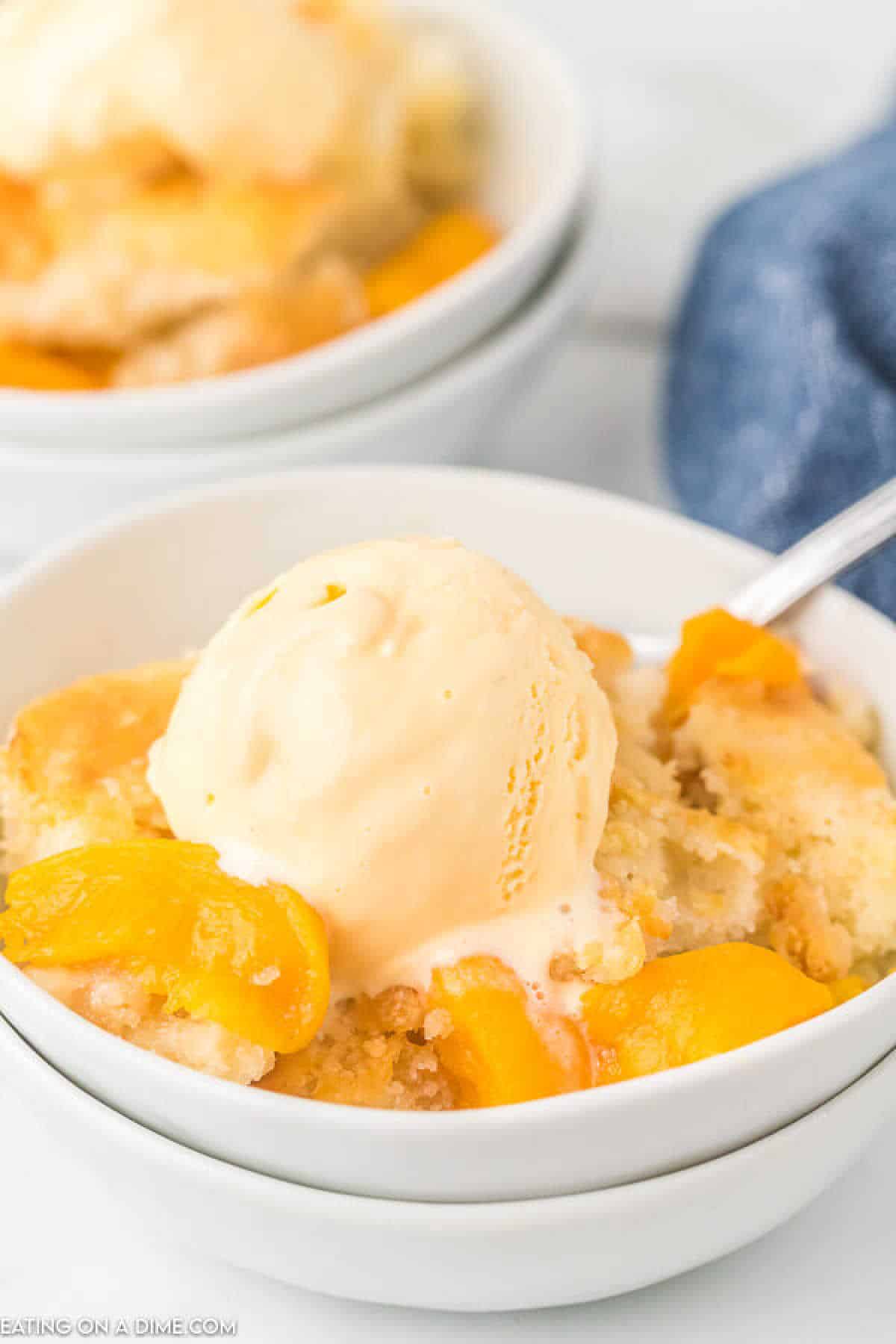 A bowl of crock pot peach cobbler with a scoop of vanilla ice cream melting on top. The dessert features golden peaches and a crumbly topping, served in a white bowl with a spoon. In the background, another bowl is partially visible.