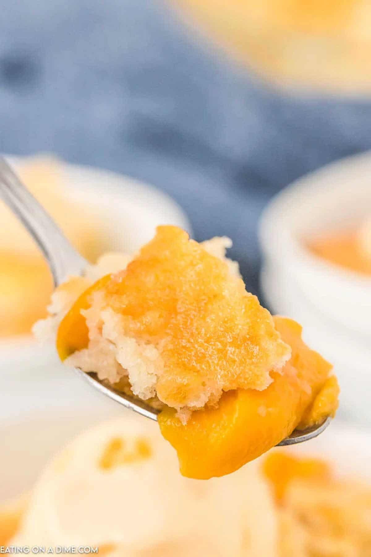 A close-up of a spoon holding a piece of peach cobbler with a golden, crumbly topping reveals the delicious results of the crock pot peach cobbler recipe. In the background, a blurred view of a bowl holds more delightful servings.