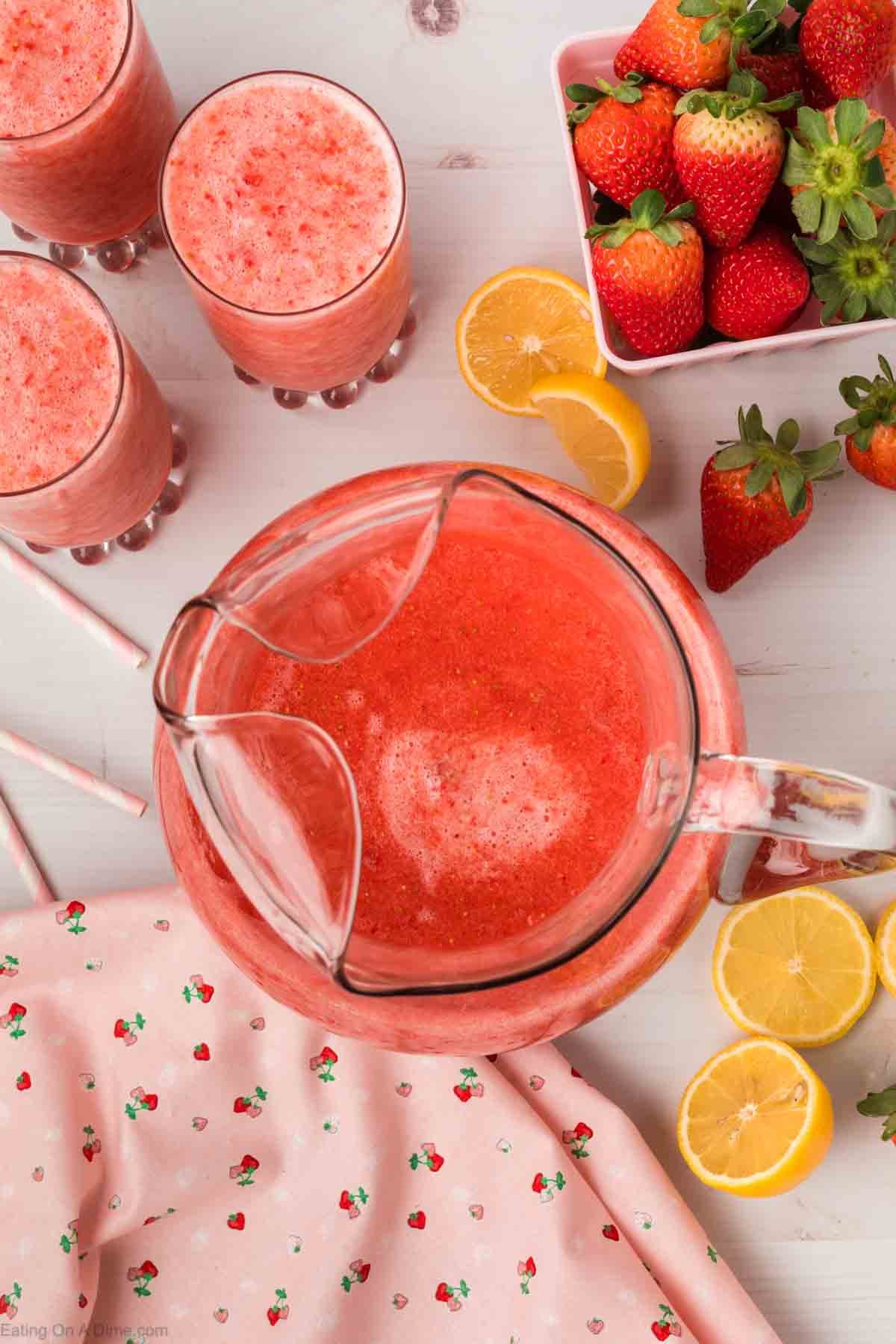 Strawberry Slushie in a glass pitcher with three glasses of strawberry slushie on the side and a basket of fresh strawberries on the side