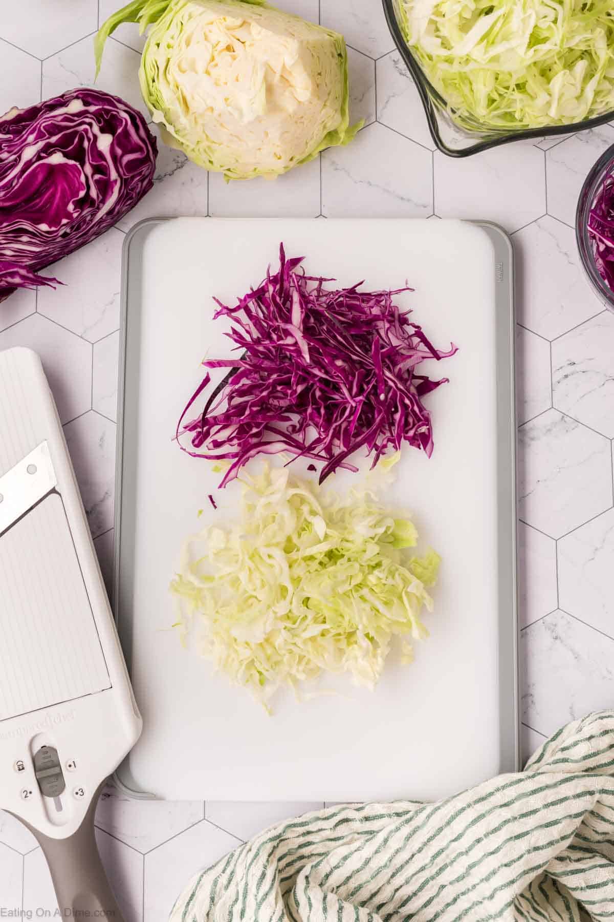 Shredded red and white cabbage on a cutting board with a mandoline slicer beside it brings you one step closer to the best coleslaw recipe. Other cabbage pieces are placed in bowls, loosely arranged on a hexagonal-patterned countertop. A striped cloth is partially visible.