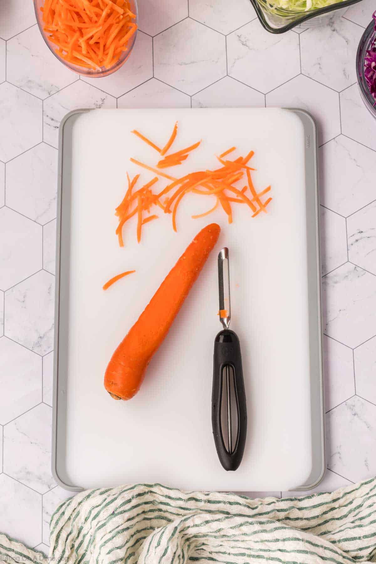 A partially peeled carrot on a white cutting board next to a peeler, hinting at the beginnings of the best coleslaw recipe. Shredded carrot pieces are scattered about, with a striped cloth draped at the bottom and bowls of shredded vegetables in the background.