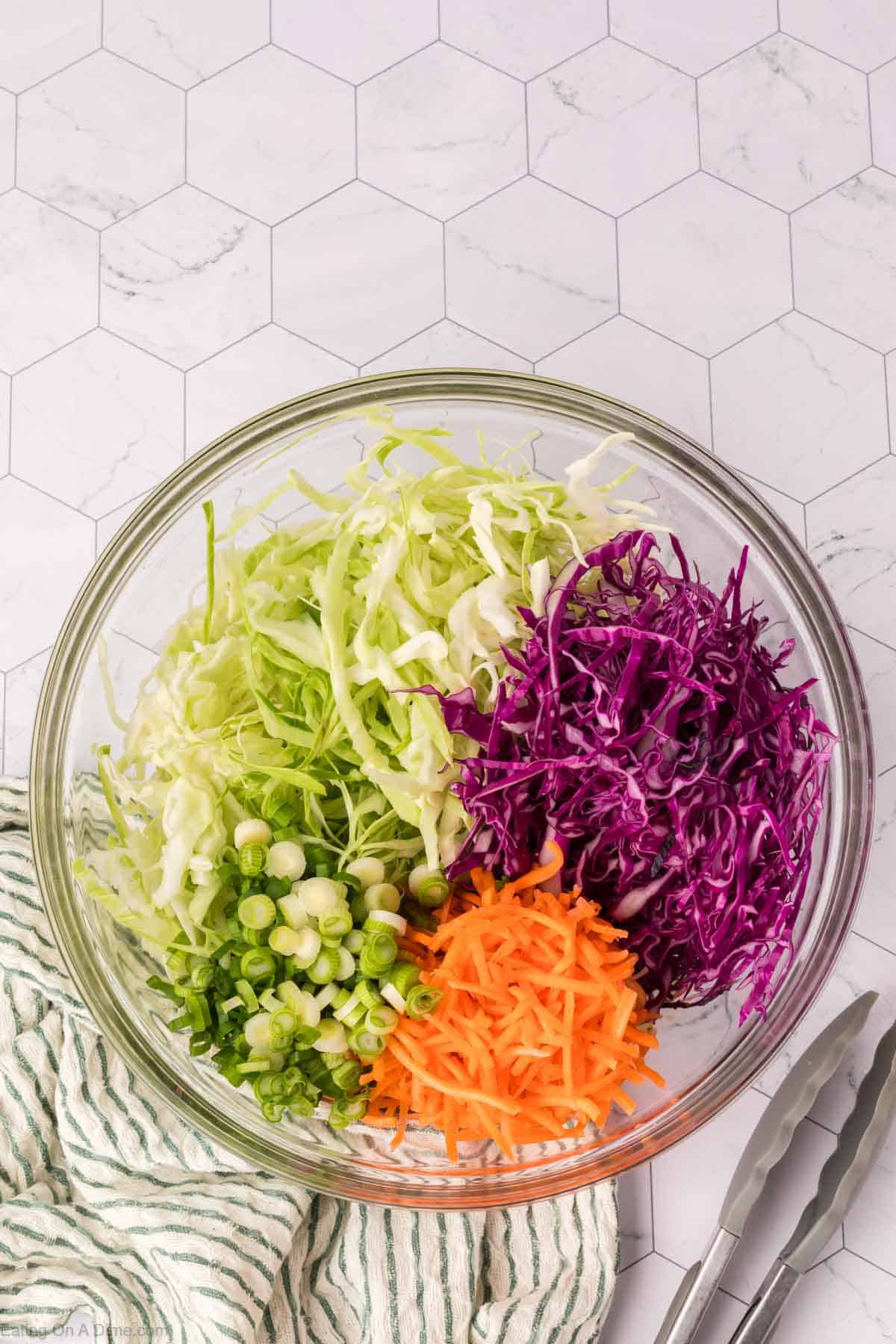 A glass bowl brimming with the best coleslaw recipe: shredded green cabbage, purple cabbage, carrots, and sliced green onions adorn a hexagon-patterned surface. A green striped cloth and metal tongs rest beside the bowl.