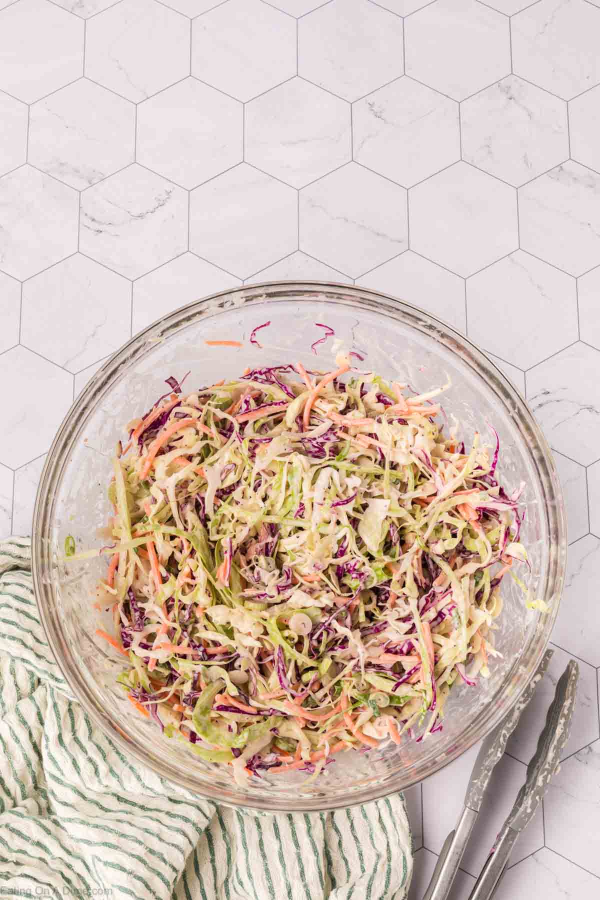 A glass bowl filled with colorful coleslaw, hailed as the best coleslaw recipe, featuring shredded cabbage, carrots, and green onions. The bowl is placed on a hexagon-tiled surface, with a green striped cloth and metal tongs nearby.