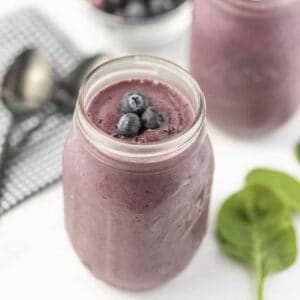 A creamy purple blueberry spinach smoothie in a mason jar, topped with fresh blueberries. In the background, there are spoons on a checkered cloth and green leaves, likely spinach, hinting at the healthy ingredients of this delicious smoothie recipe.