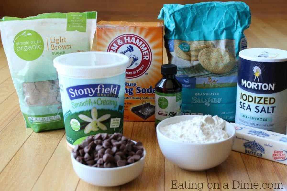 A collection of baking ingredients on a wooden surface: brown sugar, baking soda, granulated sugar, vanilla extract, salt, chocolate chips for cookie dough frozen yogurt, flour in a bowl, and butter sticks.