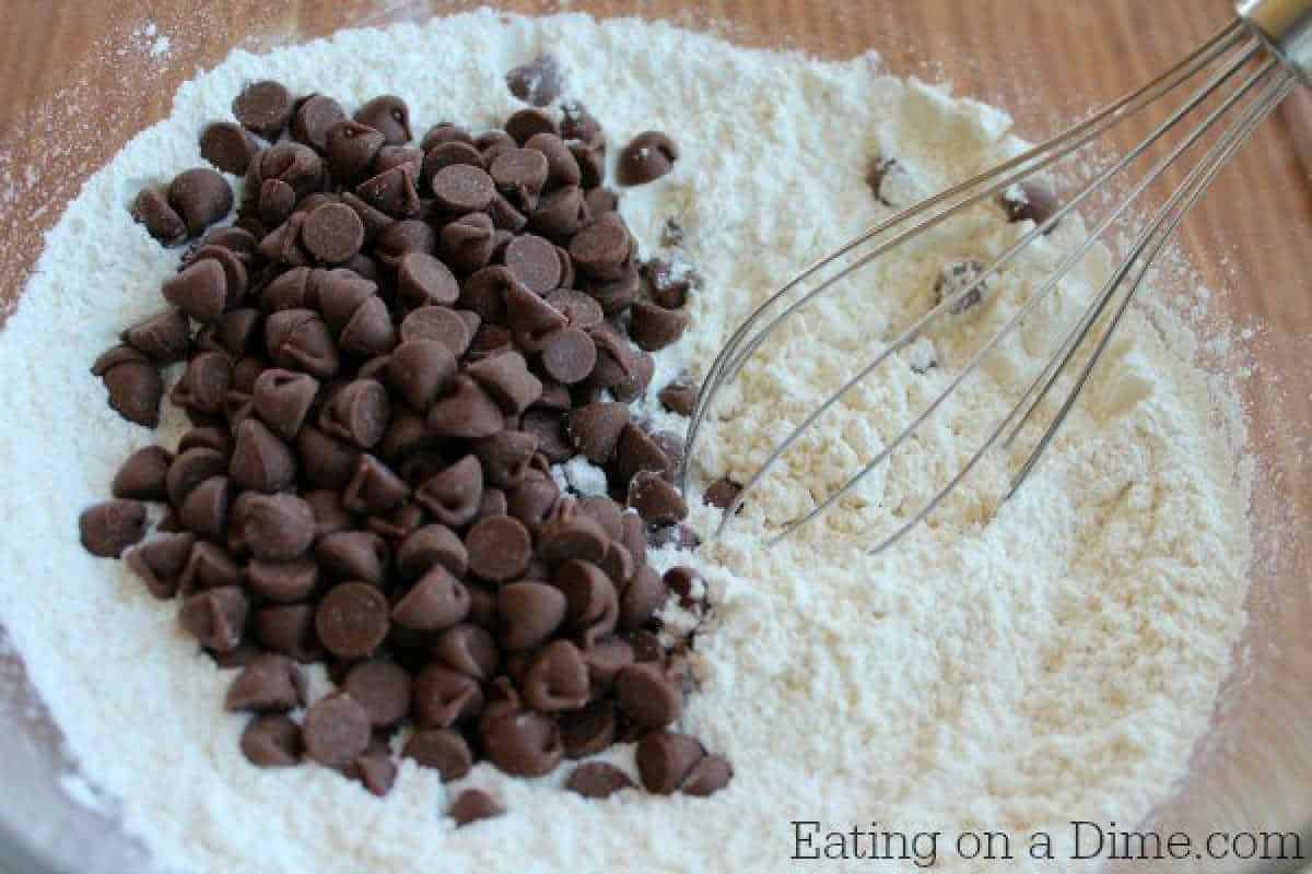 A mixing bowl with chocolate chips placed on top of a flour mixture, reminiscent of starting a delicious chocolate chip cookie dough recipe. A metal whisk rests in the bowl, ready to blend the flavors. Wooden surface underneath adds warmth to this cozy scene.