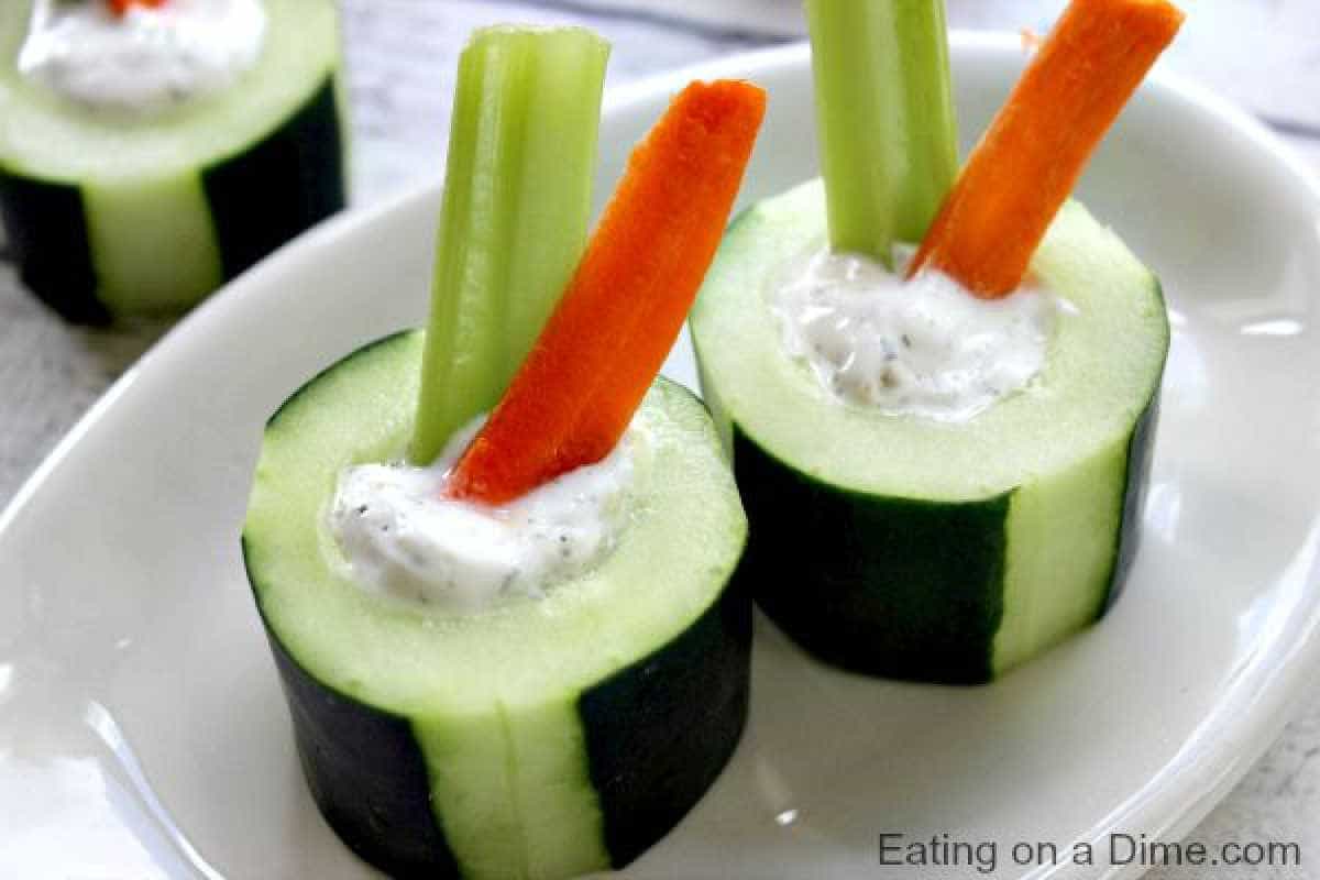 Cucumber bites, filled with creamy dip and topped with a celery and carrot stick, are elegantly arranged on a white plate.