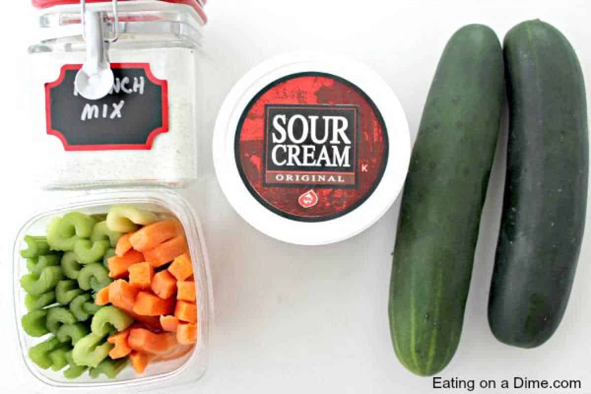 Top view of ingredients: a jar labeled "Ranch mix," a tub of sour cream, a container with chopped celery and carrots, two whole cucumbers perfect for cucumber bites.