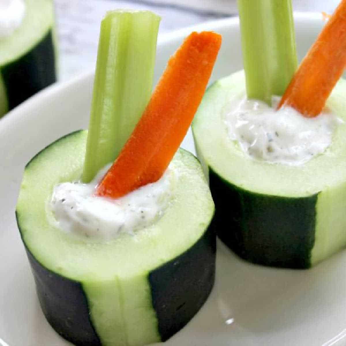 Cucumber bites are hollowed-out cucumber slices filled with creamy white dip, each cradling a celery stick and a carrot stick, elegantly arranged on a white plate.