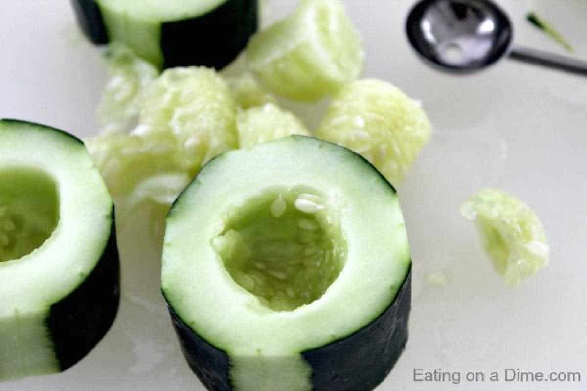 Close-up of a peeled cucumber with the seeds scooped out, resting on a white surface, ready to be transformed into refreshing cucumber bites. Several pieces of cucumber and a metal melon baller are nearby, hinting at a simple yet delightful culinary creation.