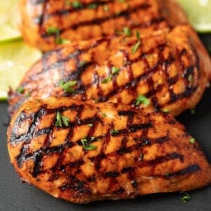 Catalina grilled chicken breasts with crosshatched grill marks, garnished with chopped herbs, elegantly placed on a dark slate surface. Lime wedges are artfully positioned in the background.