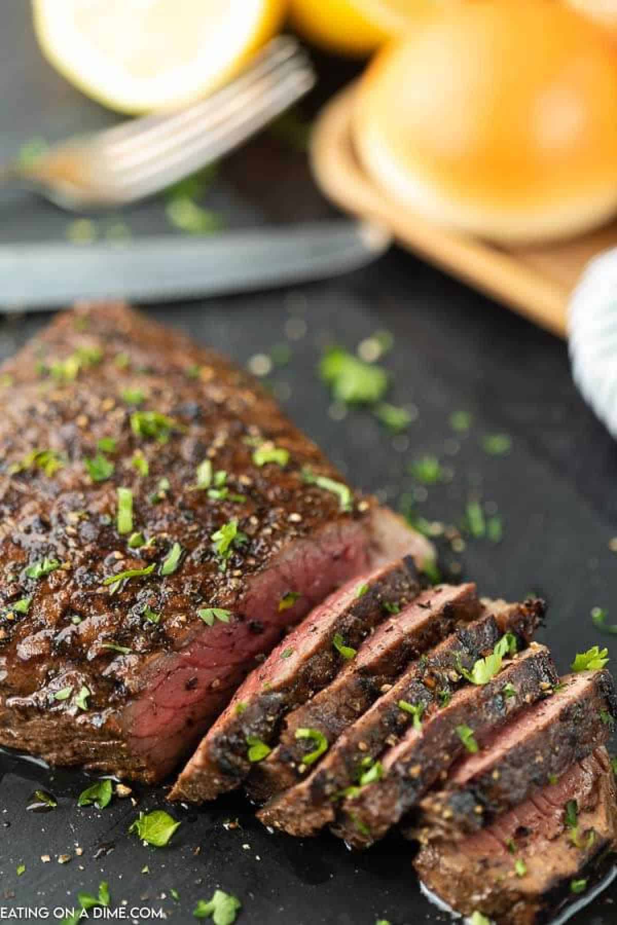 Sliced grilled steak, marinated in a zesty London broil marinade, is garnished with chopped parsley on a black surface. In the background, a fork, knife, lemon halves, and sandwich buns rest on a wooden board.