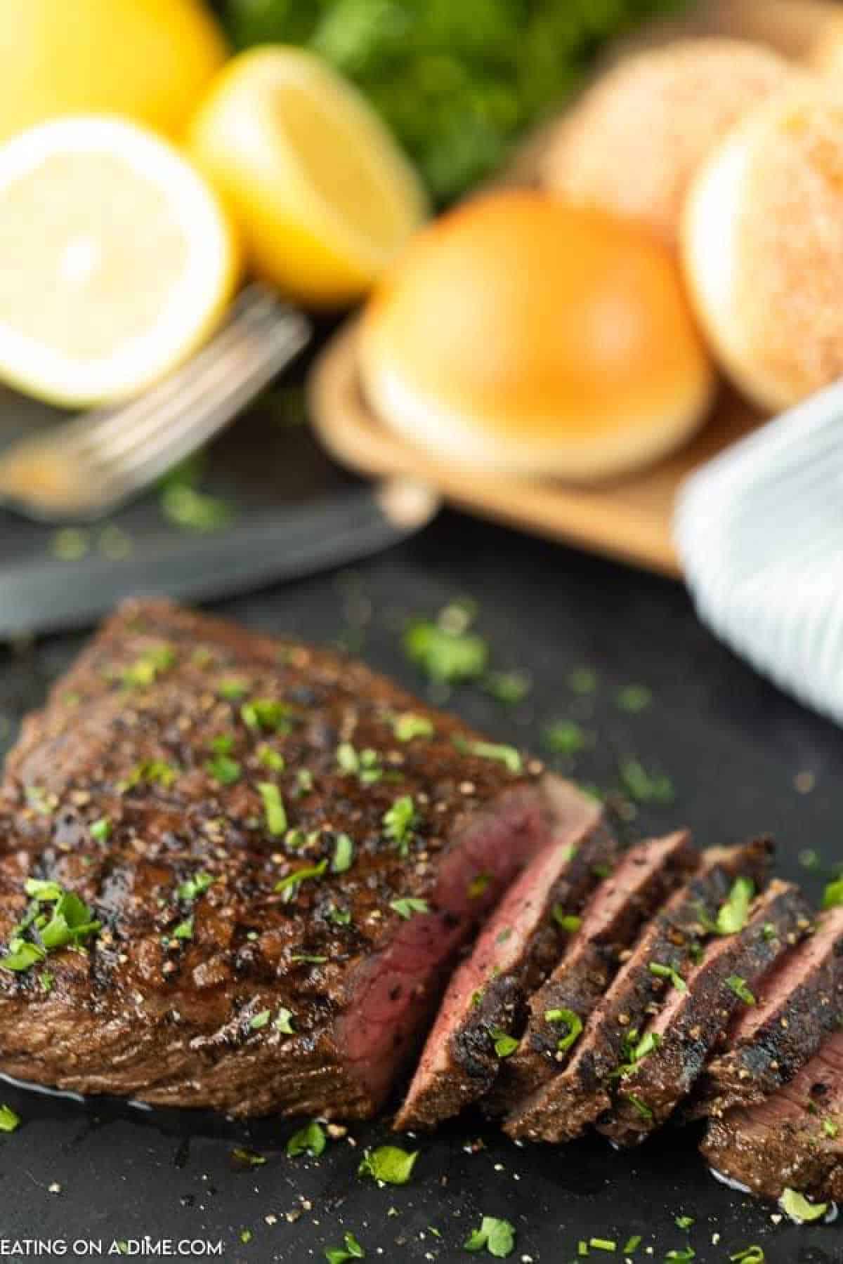 Sliced, seared steak in a rich London broil marinade is garnished with chopped herbs on a dark plate. Lemon halves, lettuce, buns, and utensils linger invitingly in the background.